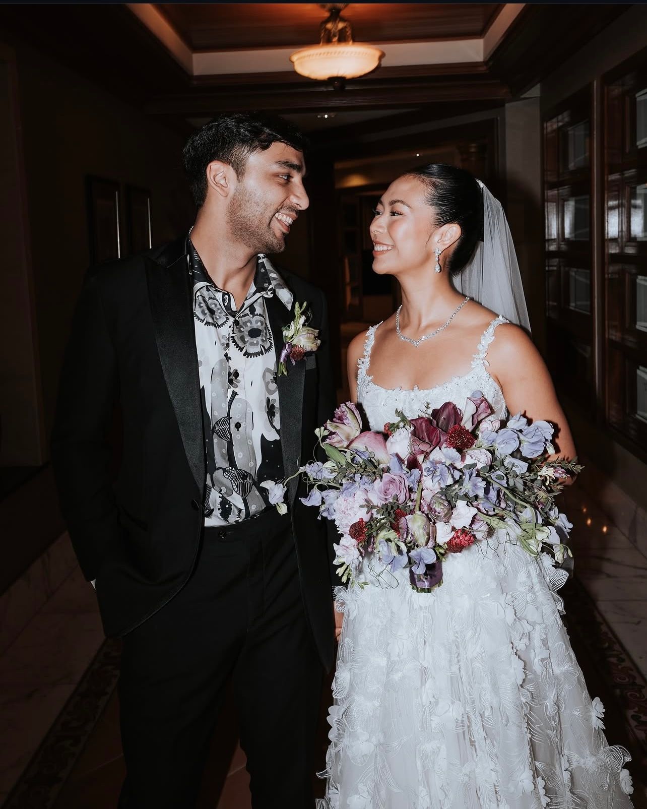 A smiling bride in a white floral-appliqué gown and veil holds a pink and lavender bouquet while laughing with her groom in a black tuxedo.
