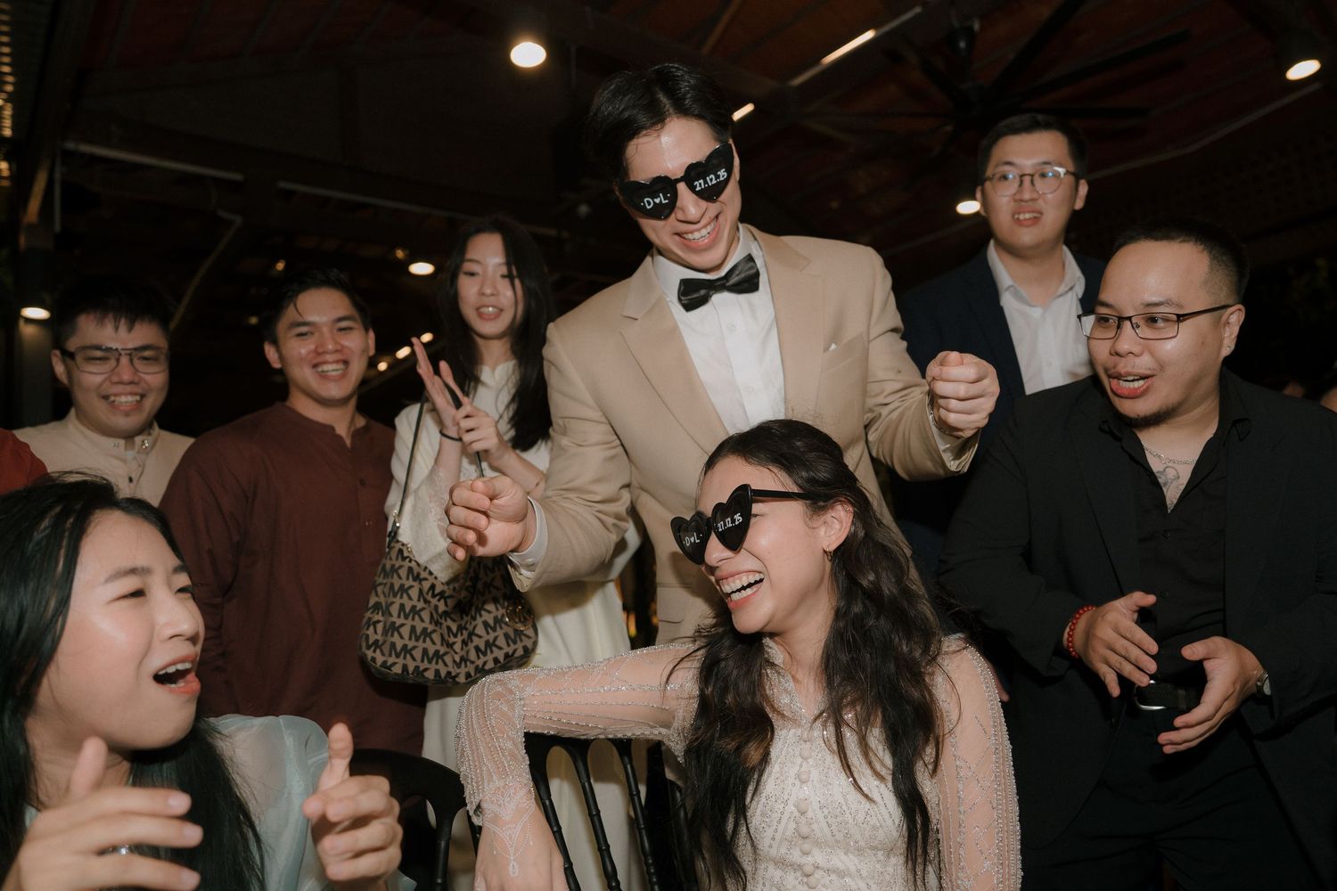 Smiling man in sunglasses, tan blazer and bow tie holds hands with a woman in sunglasses while cheerful guests watch at an indoor party.