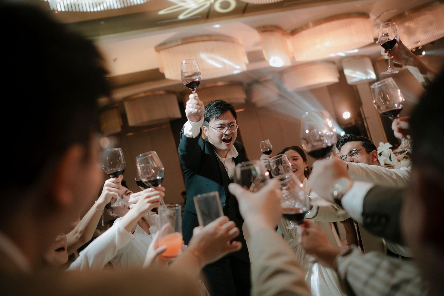 Guests raise wine glasses in a festive toast at an elegant banquet hall, with a smiling man in glasses standing at the center.