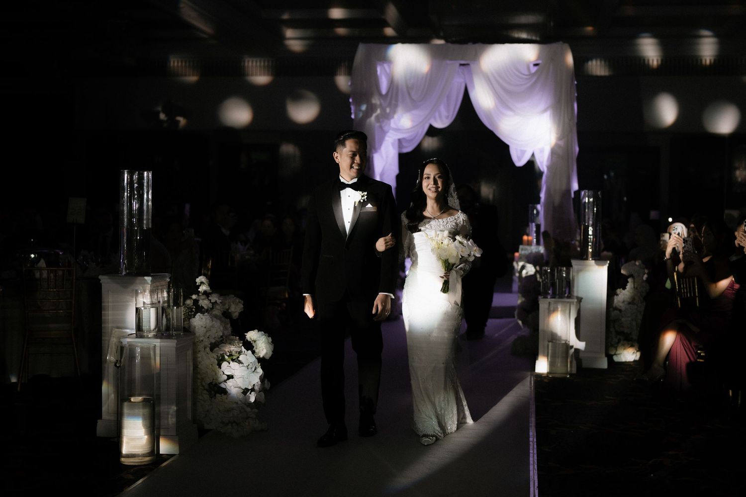 Elegant bride and groom walking down the aisle at a dark romantic wedding ceremony with white floral arch.