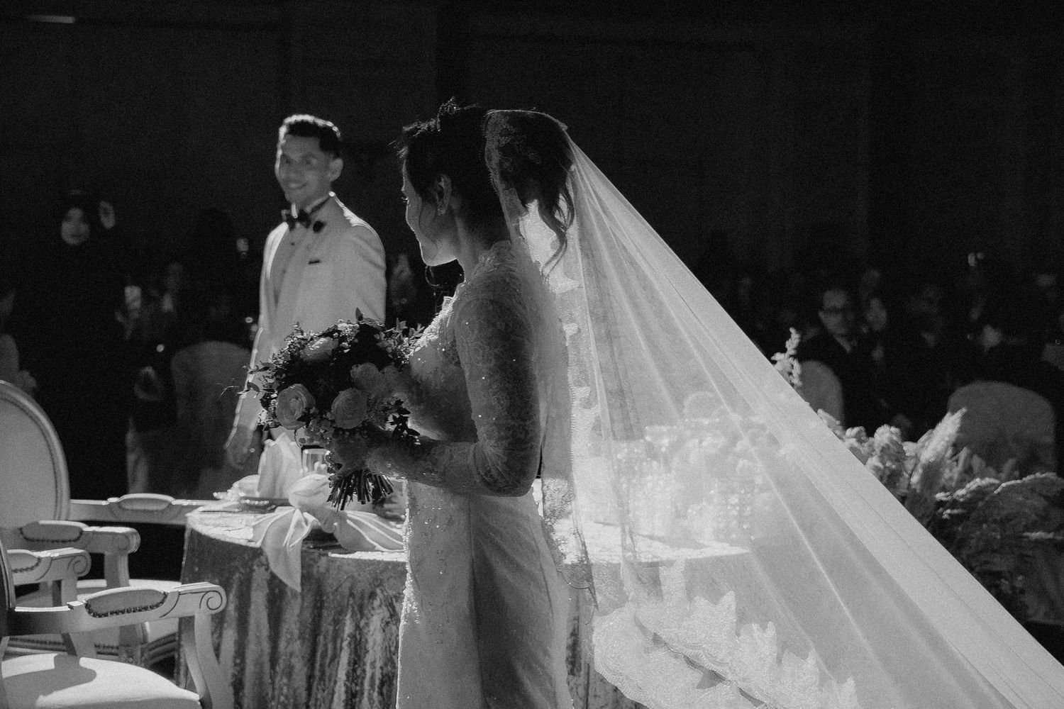 A black and white wedding reception photo showing guests toasting and celebrating at decorated dining tables.