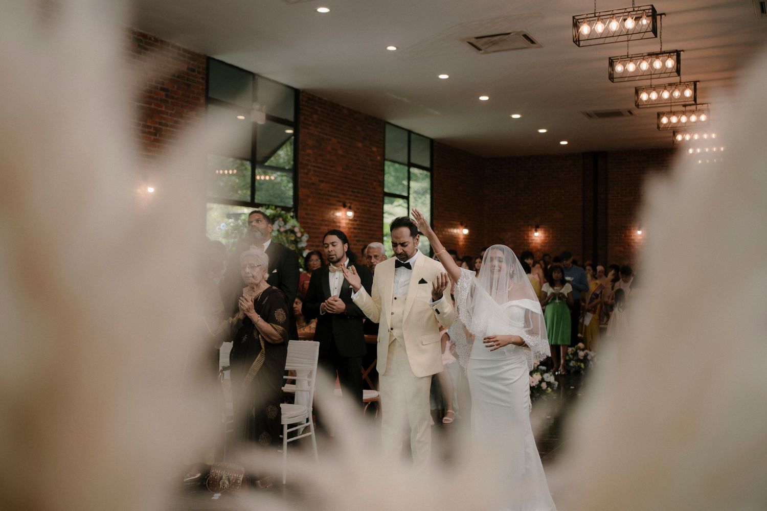 Bride in a white veil and groom in a cream tuxedo walk with raised hands as wedding guests cheer in a warm, softly lit reception hall.
