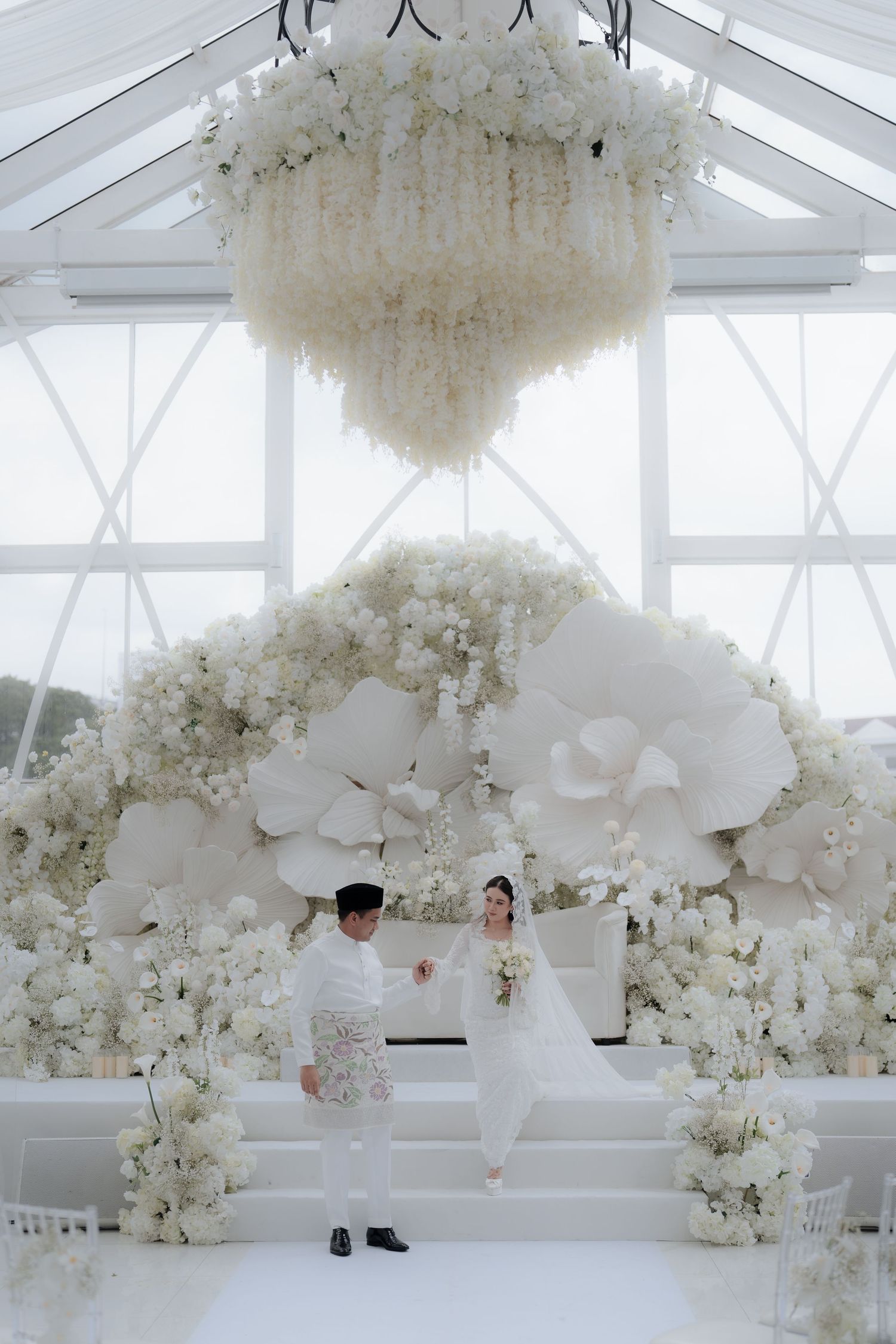 Bride in white gown and groom in traditional attire stand on steps before a giant white floral backdrop and hanging flower chandelier.