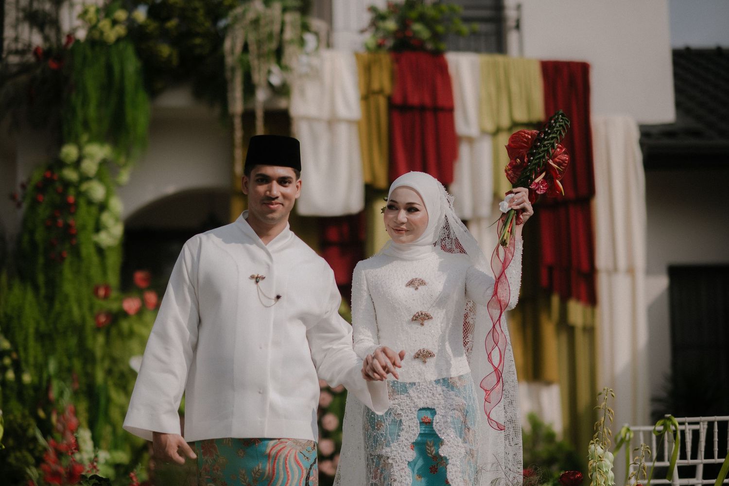 A couple in traditional Malay wedding attire, the groom in white baju melayu and bride in hijab holding a red bouquet.
