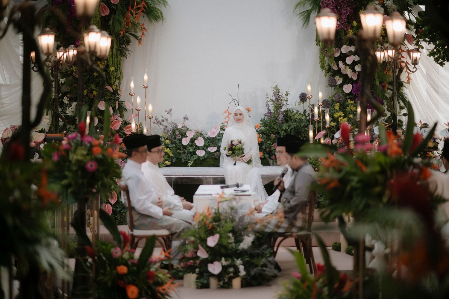 Elegant outdoor wedding ceremony with floral arch, bride in white hijab, guests seated at decorated table surrounded by lush greenery.
