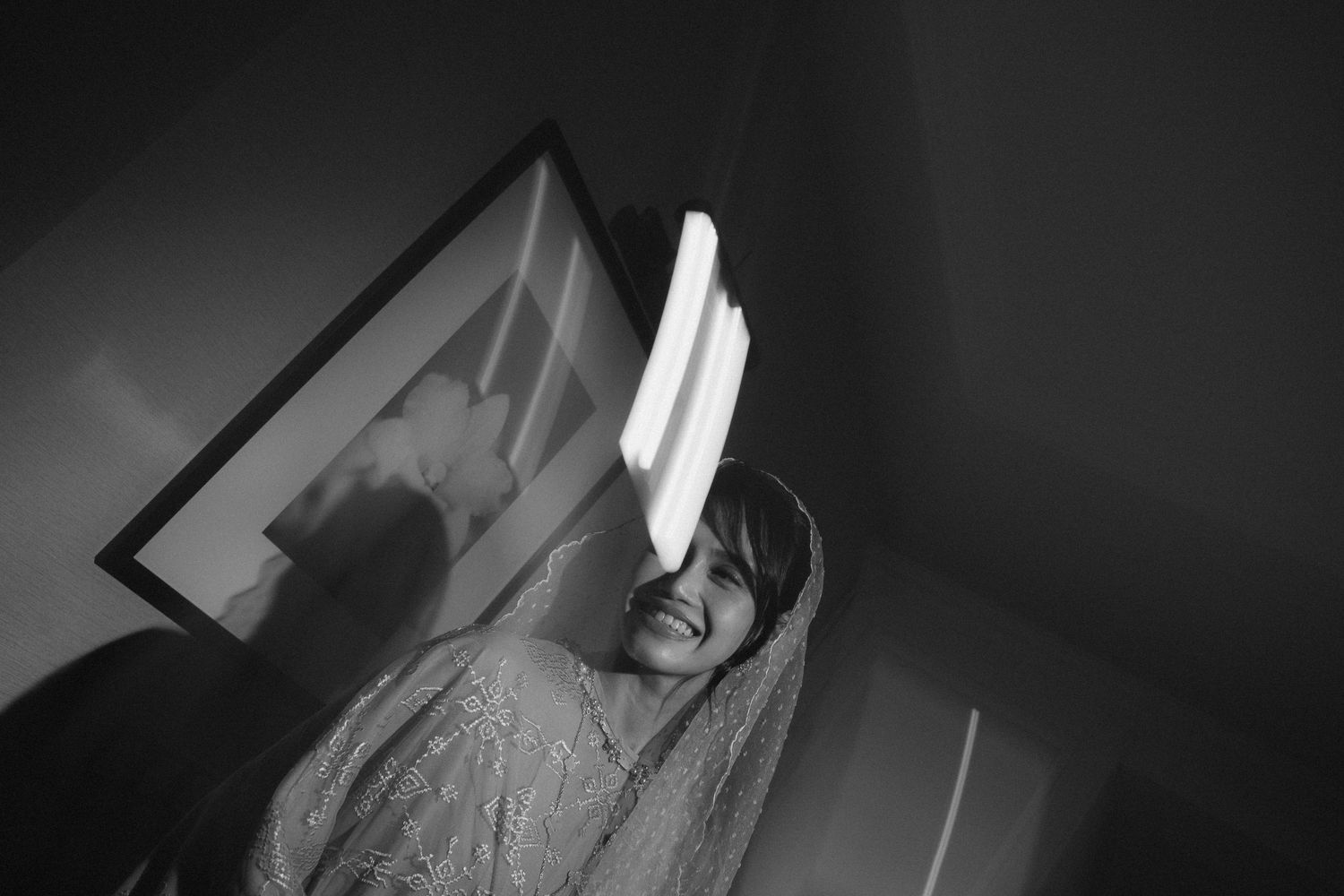 Black and white photo of a smiling bride in a veil, illuminated by a bright light, with a framed picture on the wall behind her.