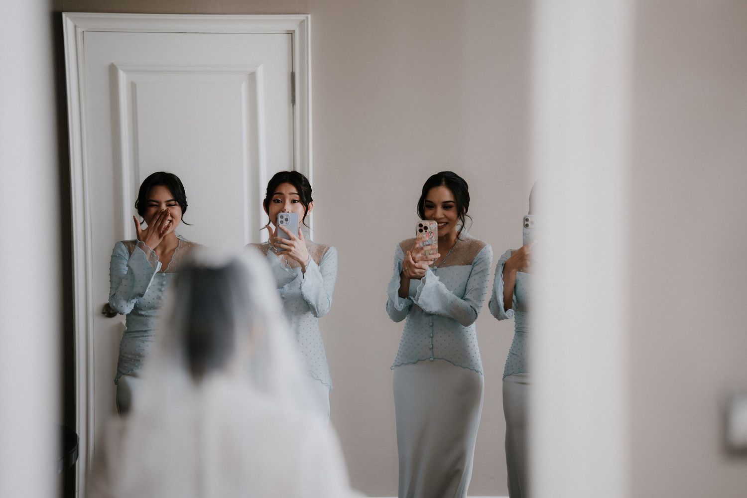 Bridesmaids in matching dusty blue dresses smile and take photos with their phones while a bride in a veil stands in the foreground.
