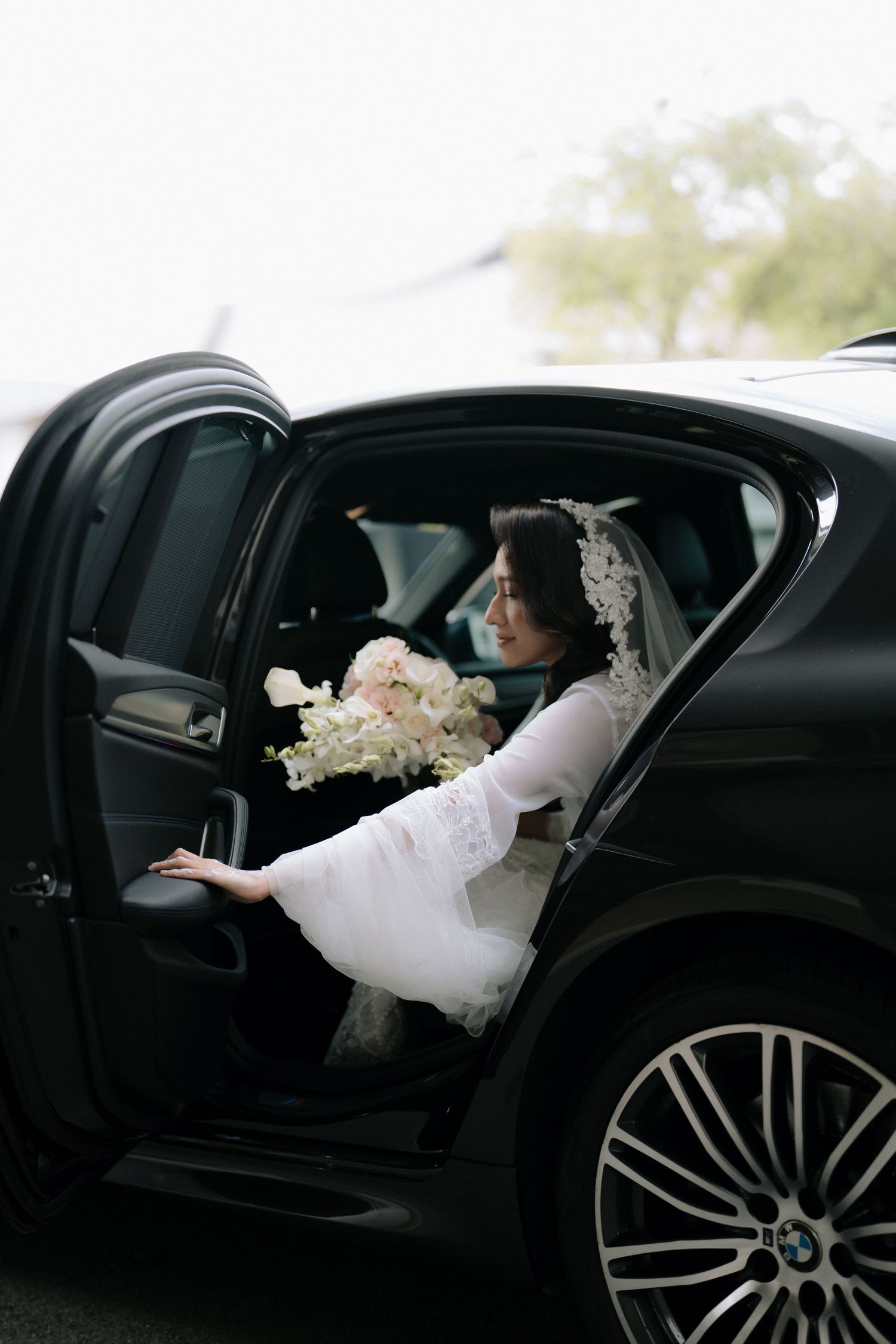 A bride in a white dress sits in a luxury black car holding a bouquet of pink and white flowers.