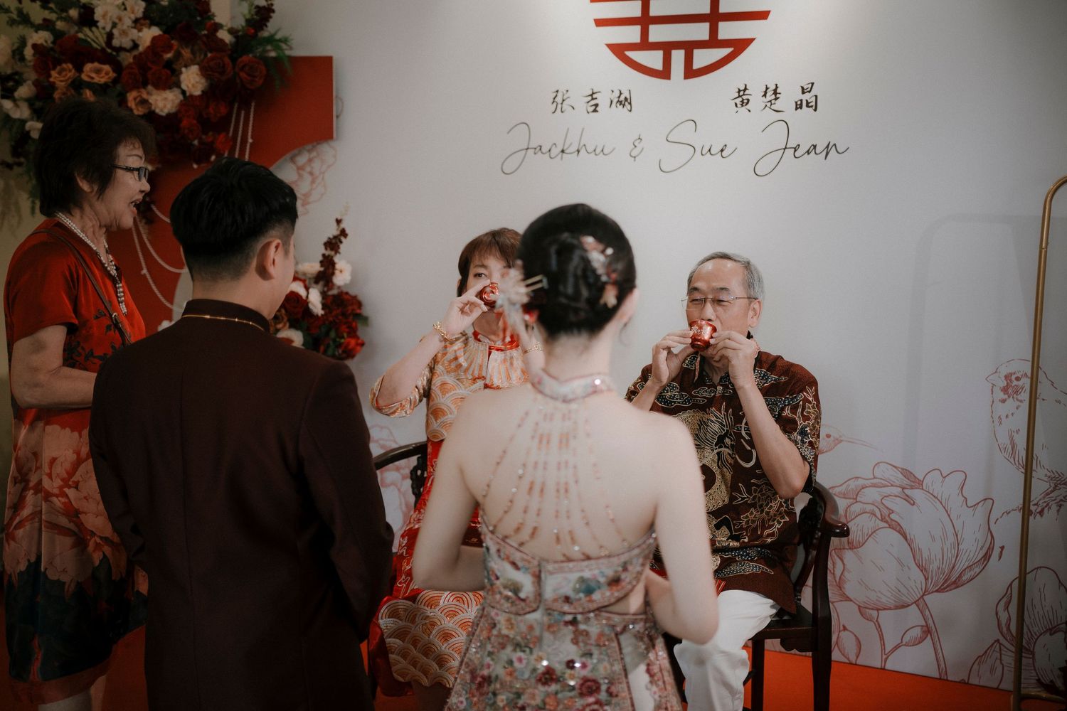 Bride in an embroidered qipao serves tea to seated elders during a traditional Chinese tea ceremony at a wedding.
