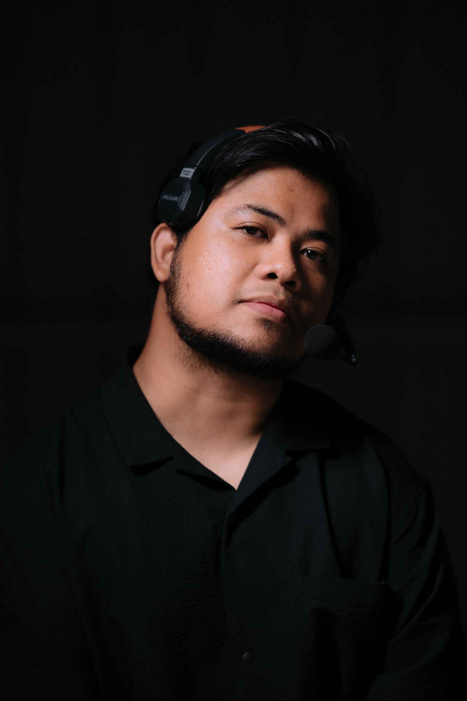 A dramatic portrait against a dark background shows a person wearing headphones and a black polo shirt in moody lighting.
