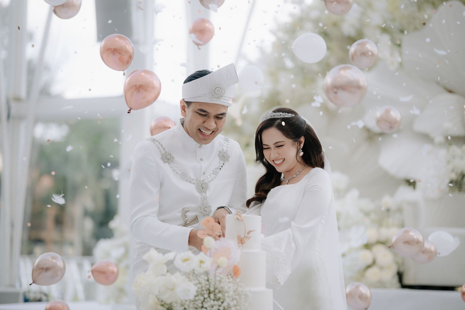 A couple in elegant white traditional wedding attire stands together under floating pink balloons and floral decorations.