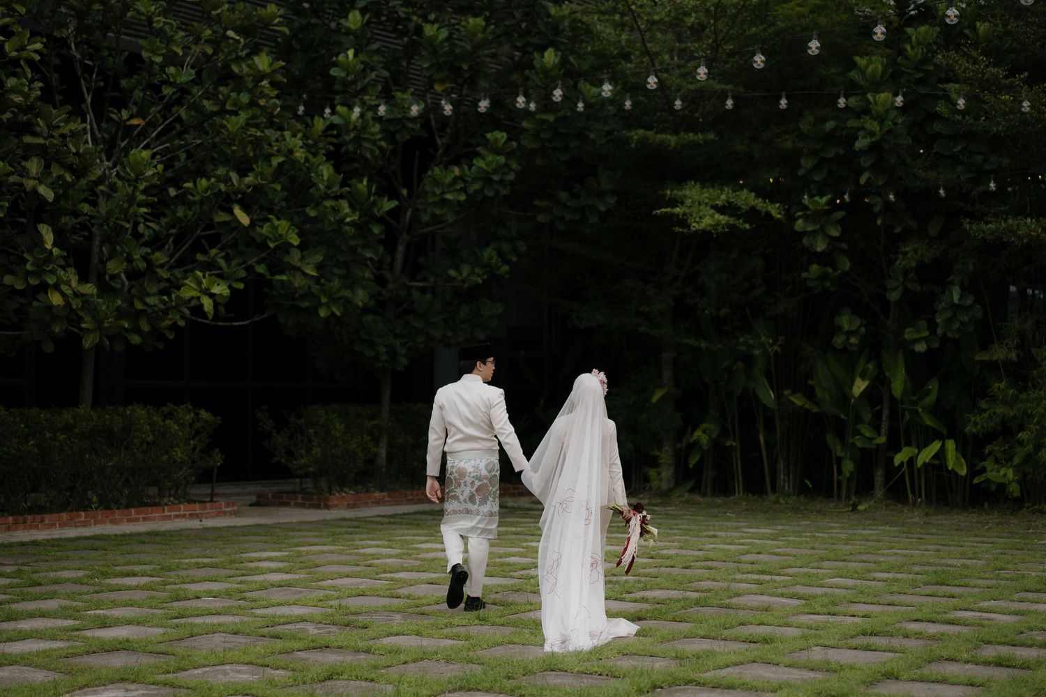 A couple strolls on a garden path with string lights overhead during an outdoor wedding at Forest Valley Malaysia