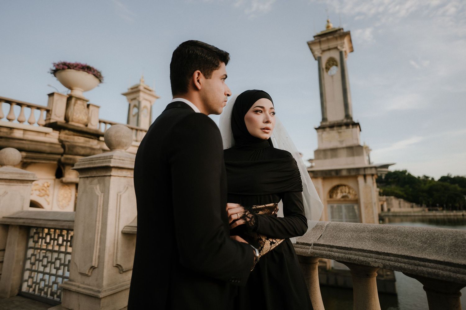 Couple in black formal attire pose on a stone balustrade bridge with a historic tower and warm sunset sky in the background.