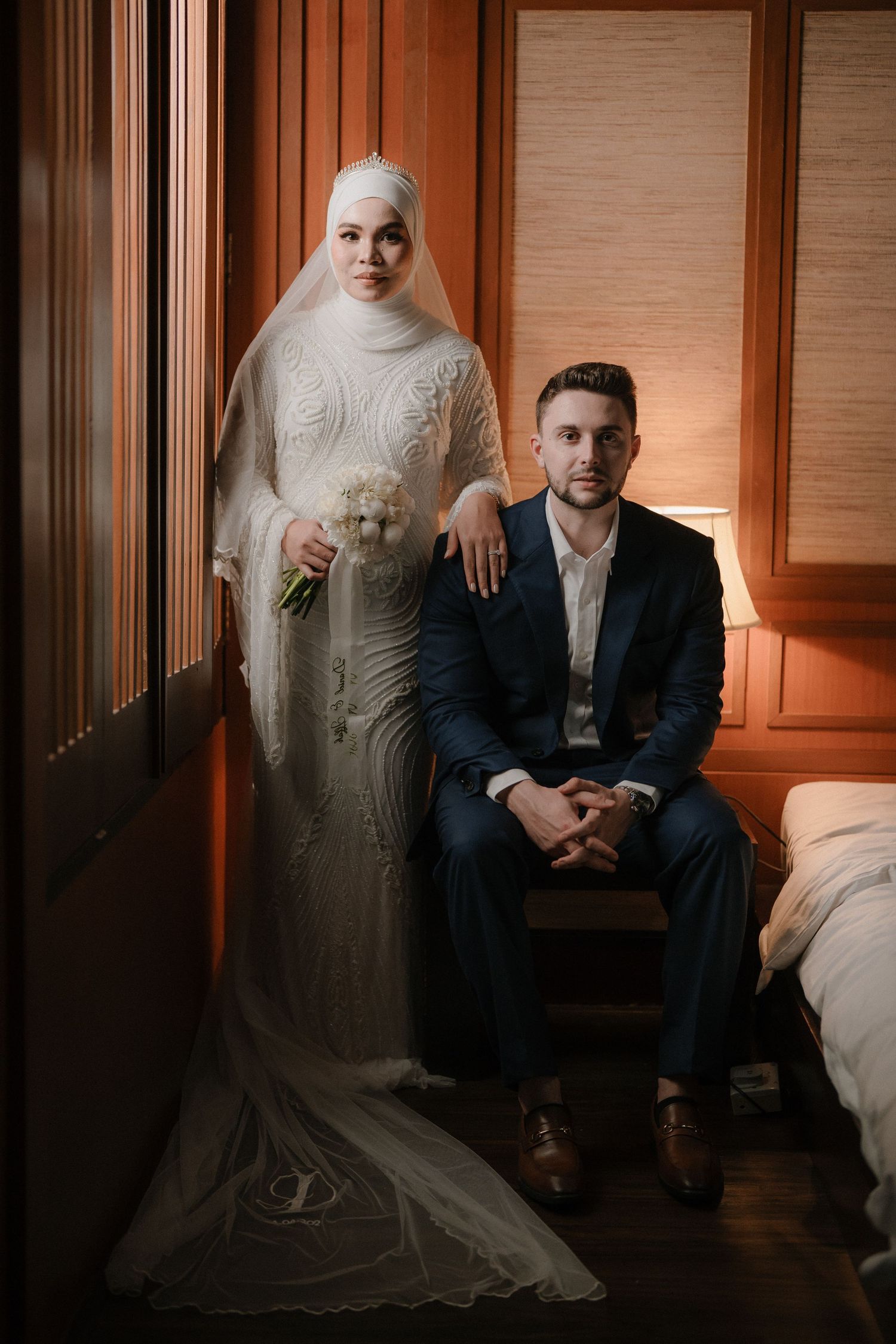 Muslim bride in white hijab gown holds bouquet beside groom in navy suit seated in warmly lit room.