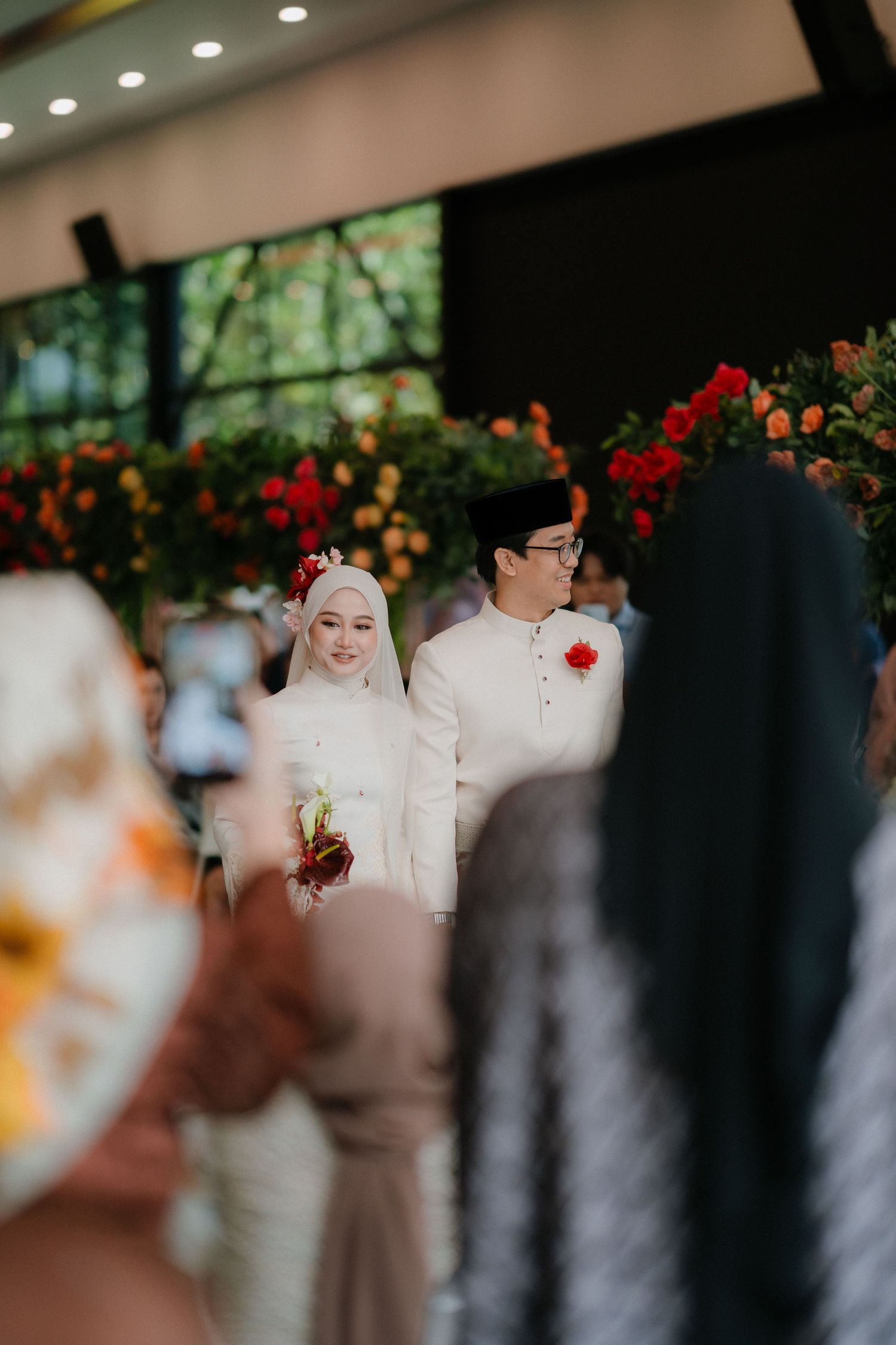 Bride in white hijab and groom in cream outfit with black songkok walk past red and orange floral decorations at a wedding as guests watch.