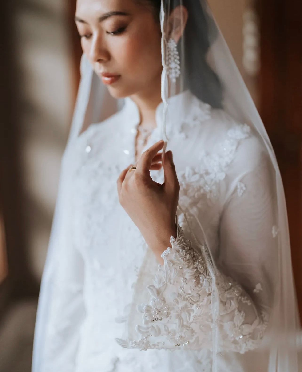 Bride in an elegant white lace gown and sheer veil gazes downward, delicately touching her hand, with sparkling drop earrings.