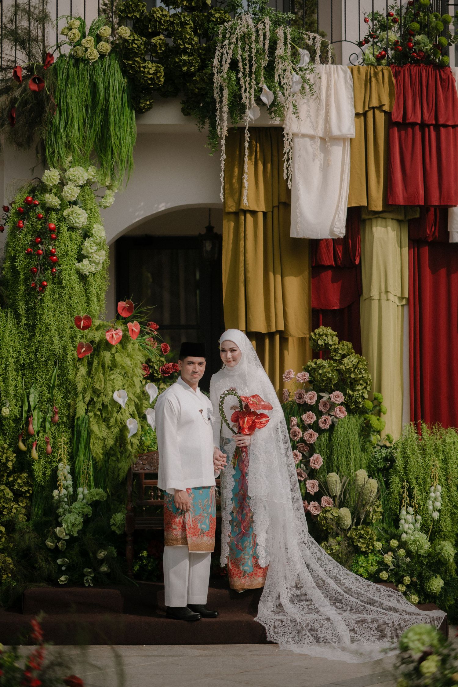 Muslim couple in traditional wedding attire posing among lush greenery and colorful floral decorations.