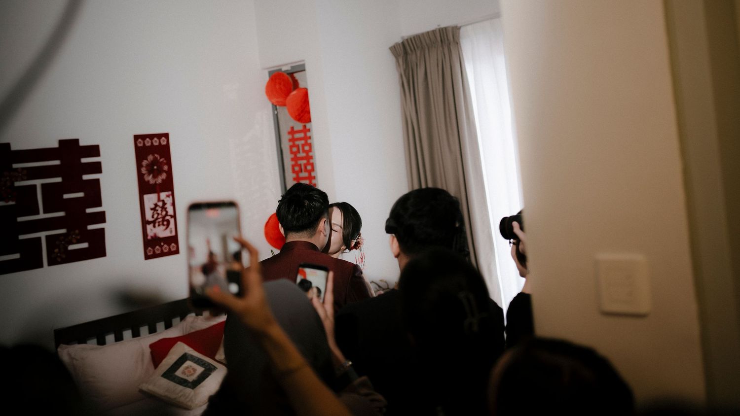 Guests gather in a room decorated with red Chinese double happiness symbols and paper decorations while someone films with a smartphone.