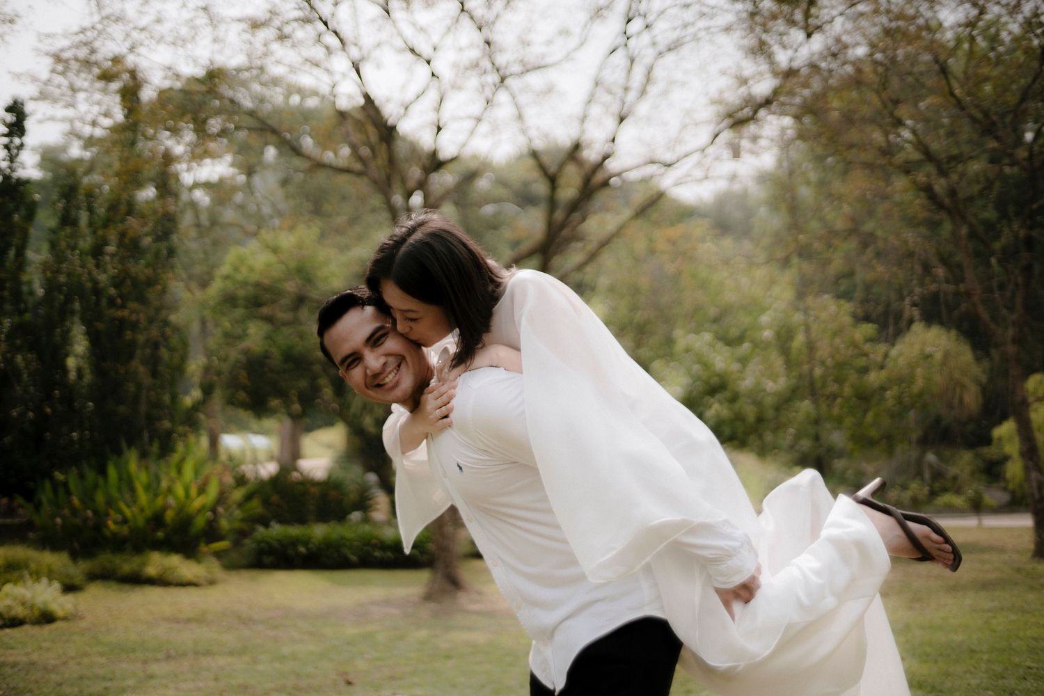 Smiling couple embracing outdoors as a man in a white shirt lifts a woman in a flowing white dress in a sunny, tree-filled park.