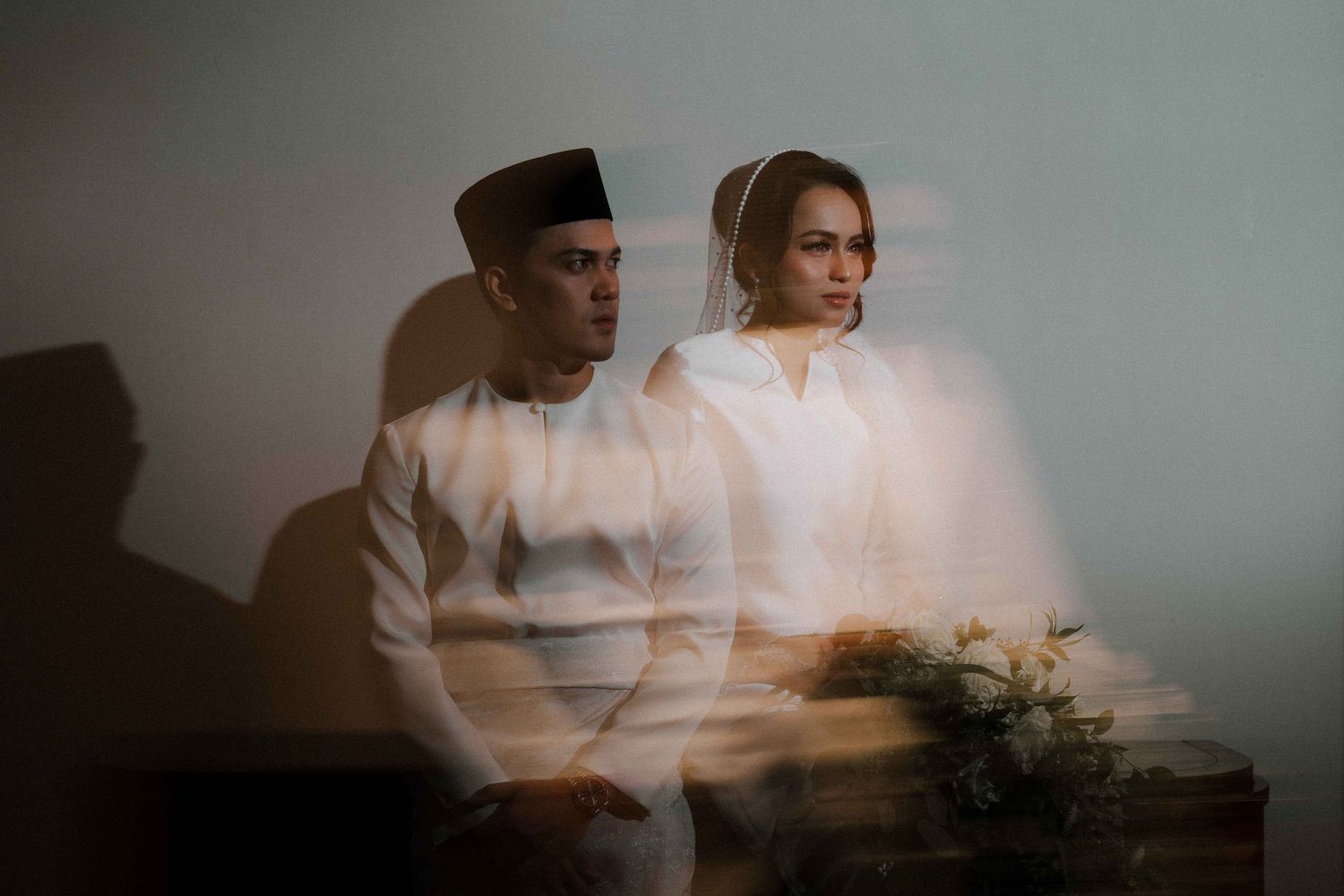 A couple sits together on a couch in soft, dreamy lighting while holding a floral bouquet.