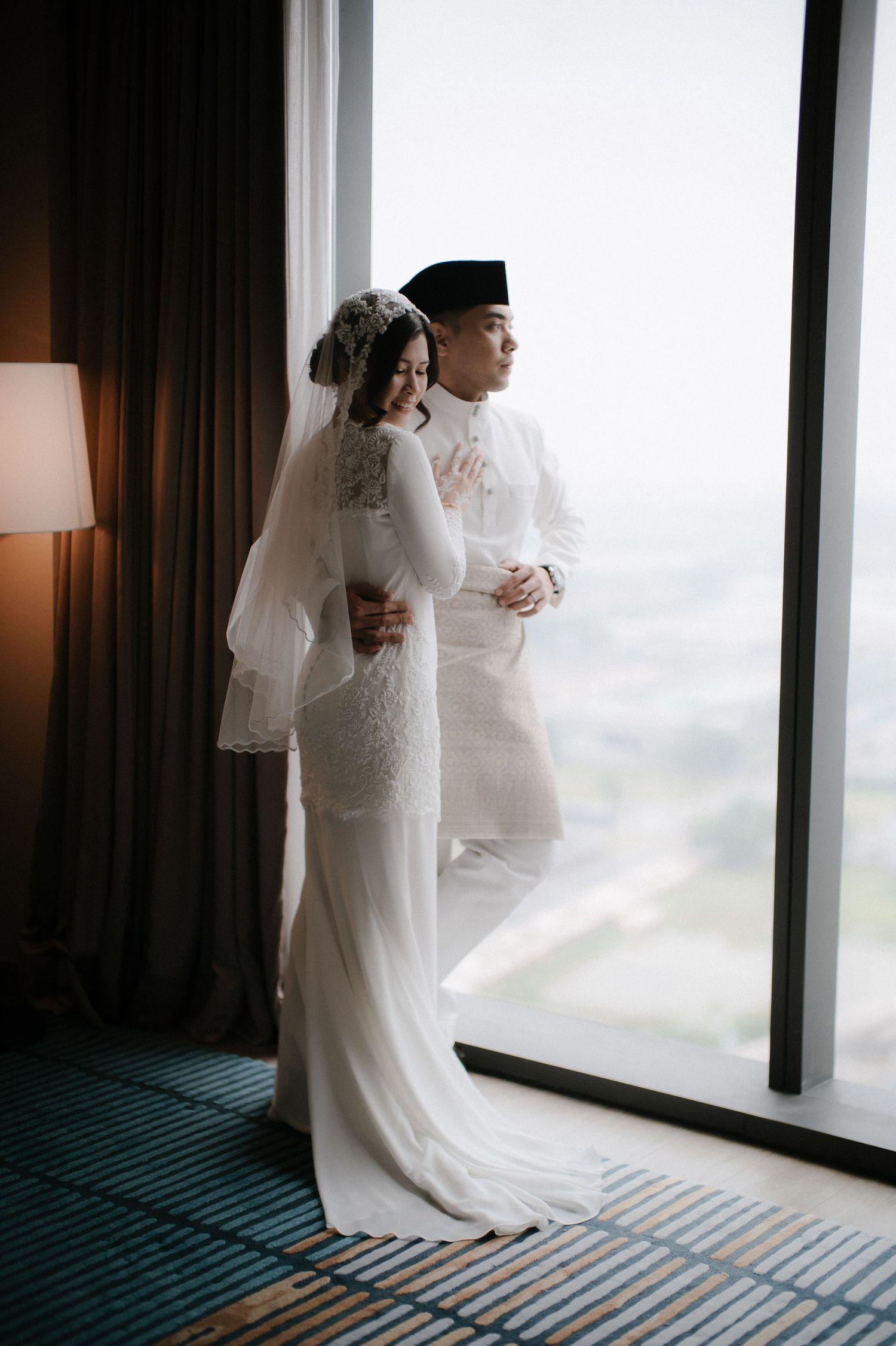 Malay bride and groom in white traditional attire standing by a large window, sharing a tender embrace on their wedding day.