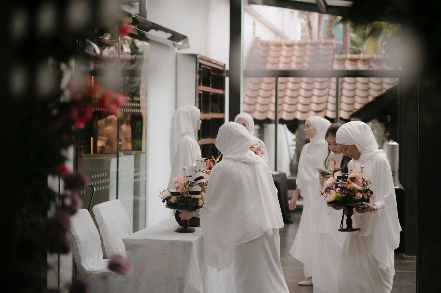Women in white hijabs and flowing white dresses stand in a bright room holding vases of pink flower arrangements beside a white table.