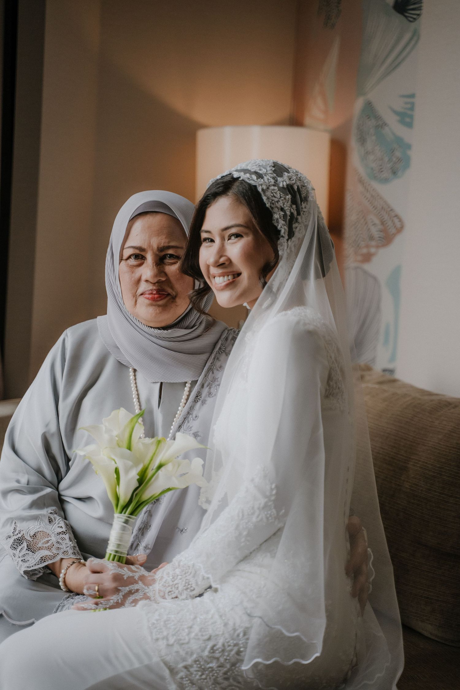 A smiling bride in a white lace gown and veil sits beside an older woman in a gray hijab, holding a bouquet of white calla lilies.