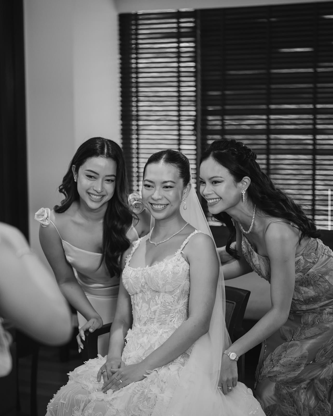 Black and white photo of a smiling bride in a lace wedding gown and veil posing with two bridesmaids in a bright room.