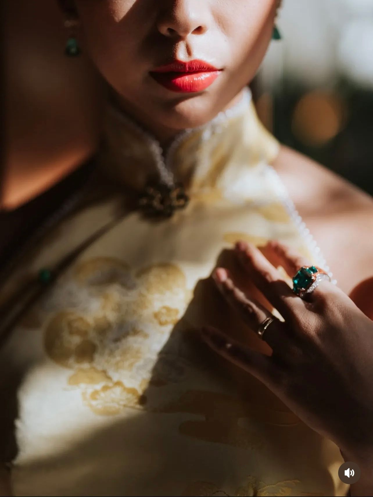 Woman in red lipstick wearing a cream qipao with gold embroidery, a jade ring and a green gemstone ring, softly lit by warm sunlight.