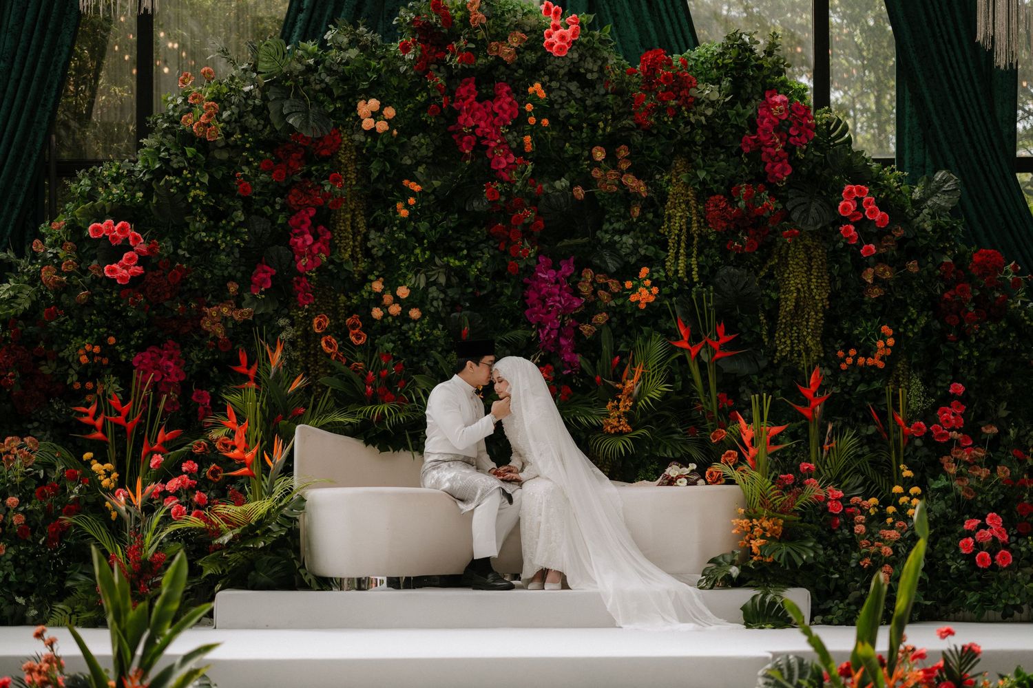 A couple shares a romantic moment in a lush garden setting with vibrant red flowers and greenery.