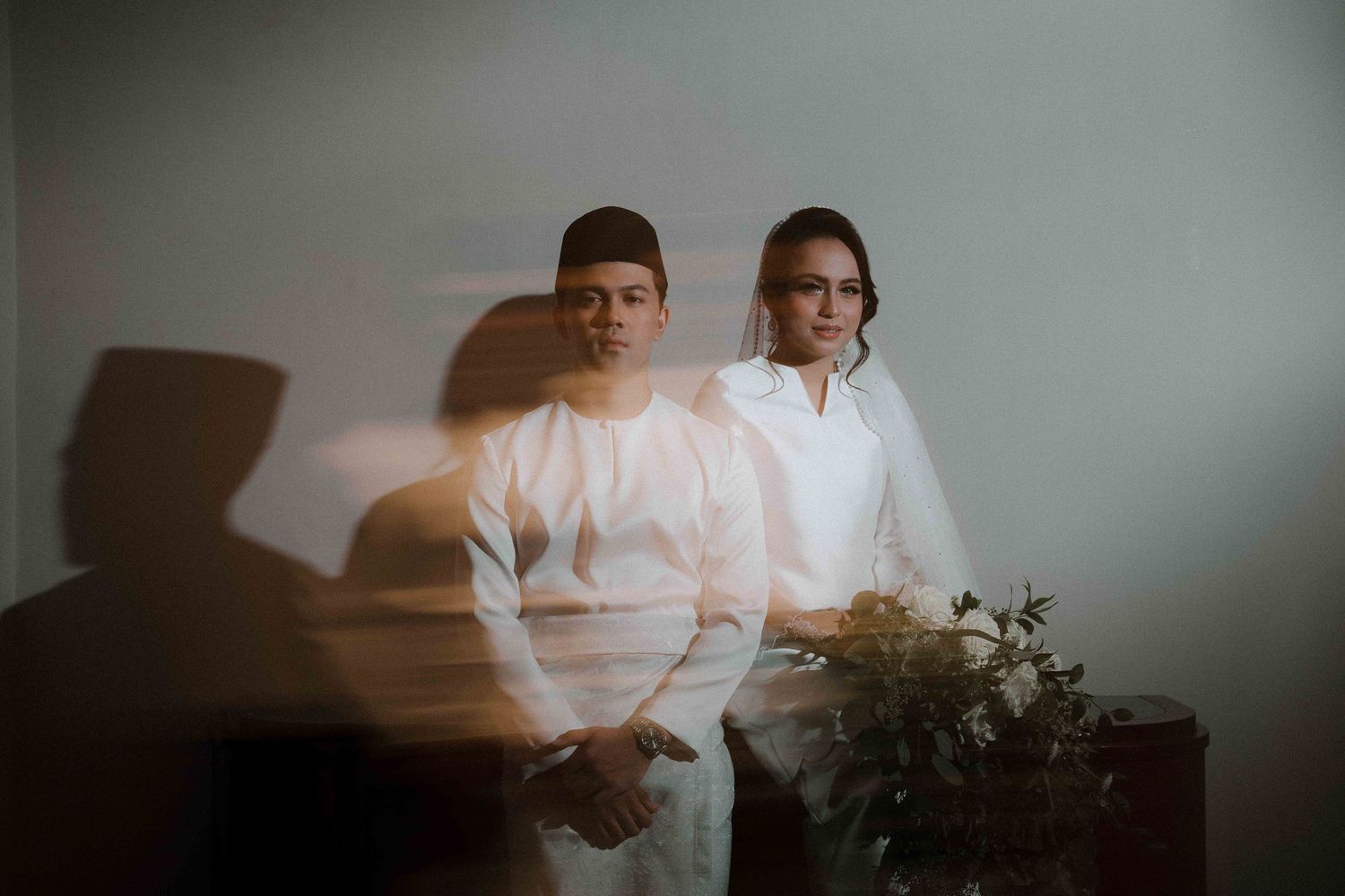 Artistic moody portrait of couple in white outfits standing against wall with dramatic shadow and plant decoration.