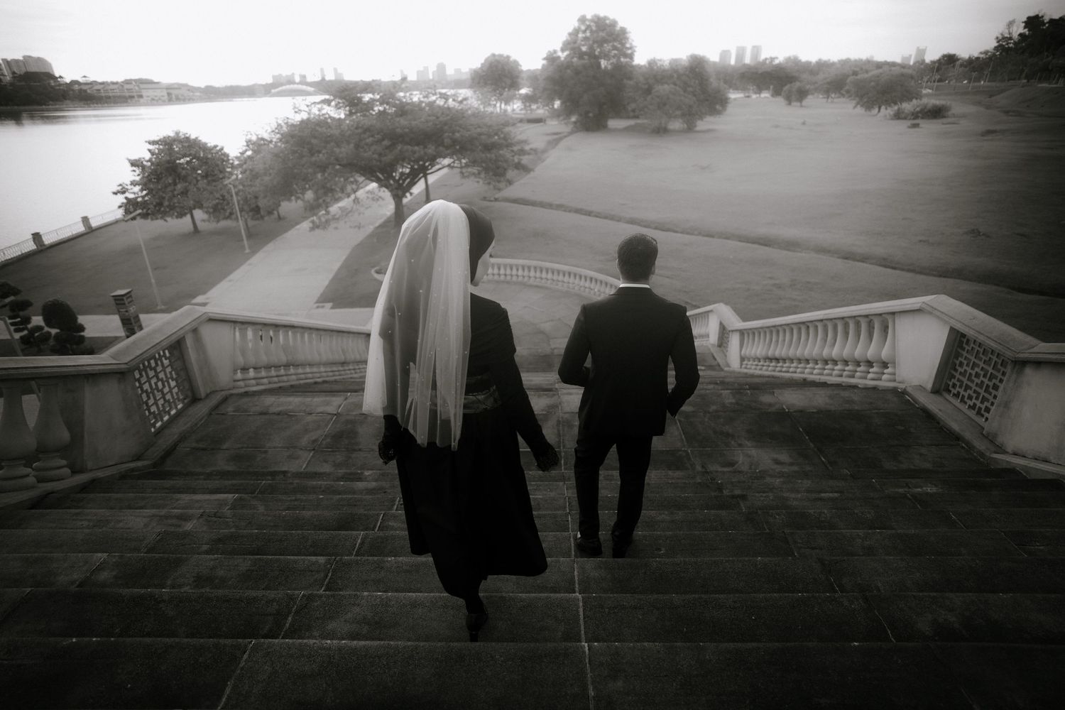 Black and white photo of a bride and groom walking up stone steps toward a scenic waterfront garden.