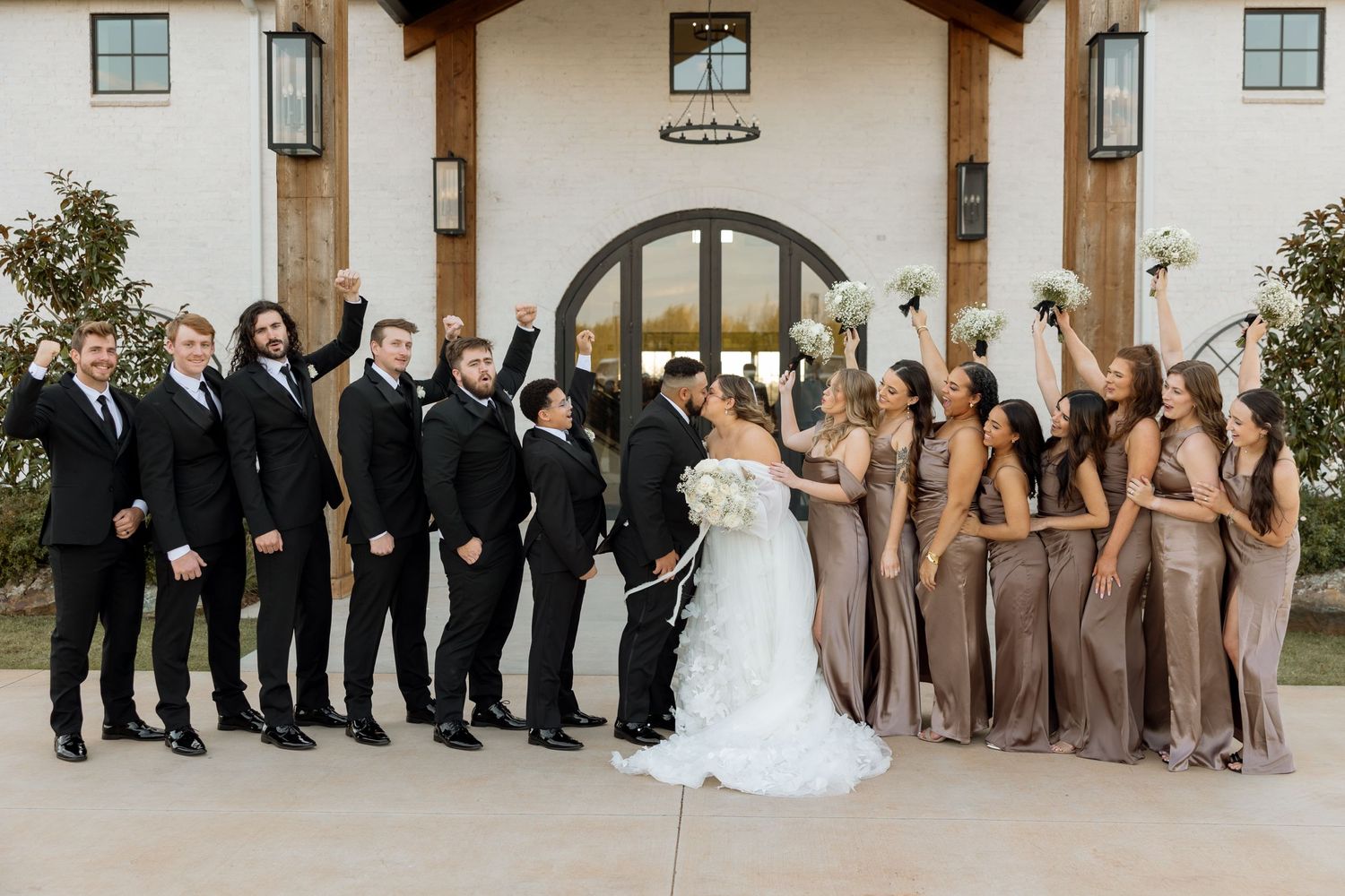 A wedding party poses outside their venue wearing black suits and mocha bridesmaid dresses.