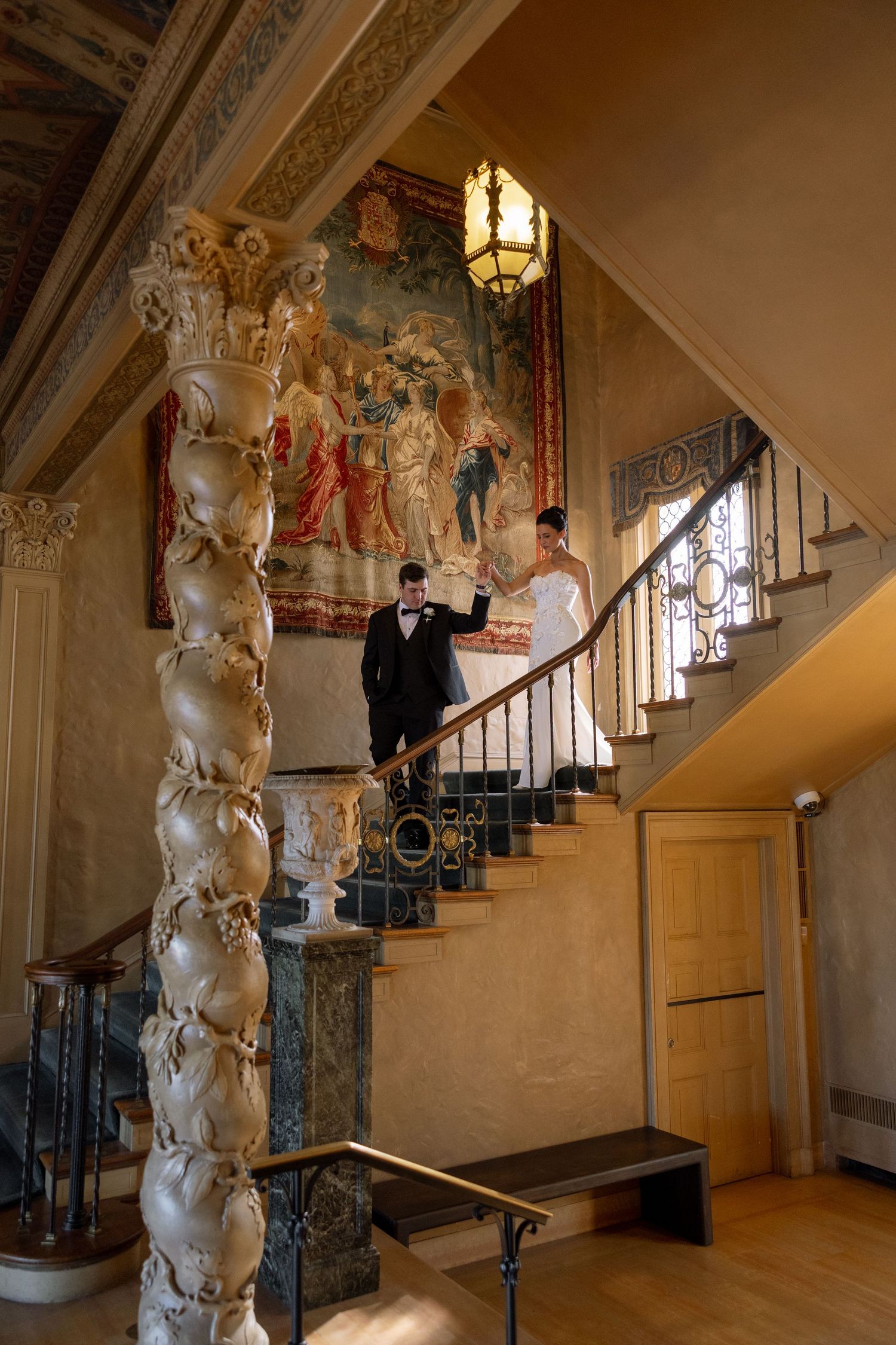 Bride and groom descend ornate staircase in historic mansion with tapestry and decorative column.