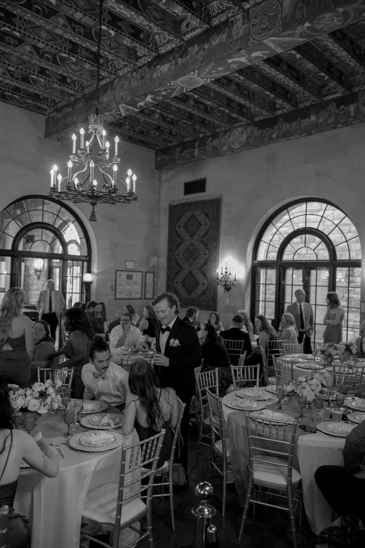 An ornate reception hall with chandeliers and arched windows where wedding guests dine at decorated tables.