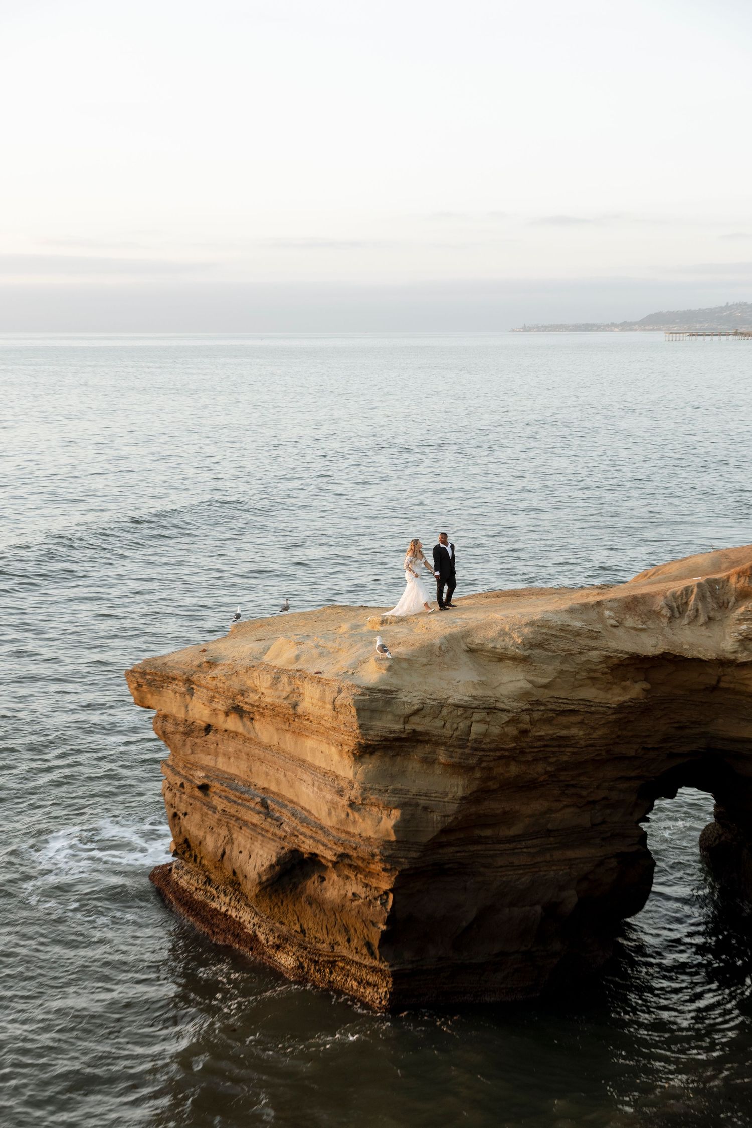 A couple stands on a dramatic ocean cliff edge overlooking the sea at sunset.