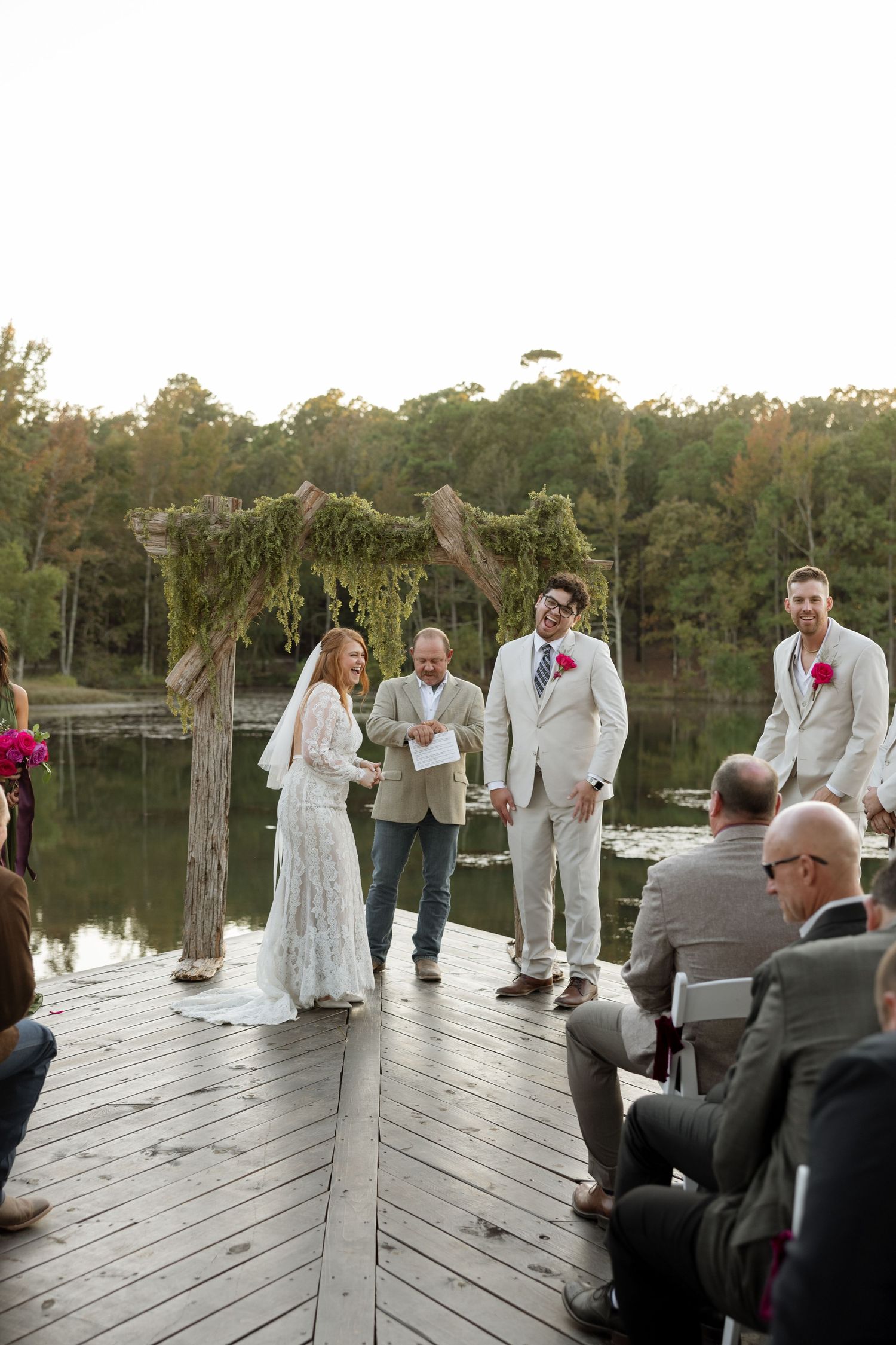 Wedding ceremony in progress on a wooden dock over a lake with a moss-covered arch.
