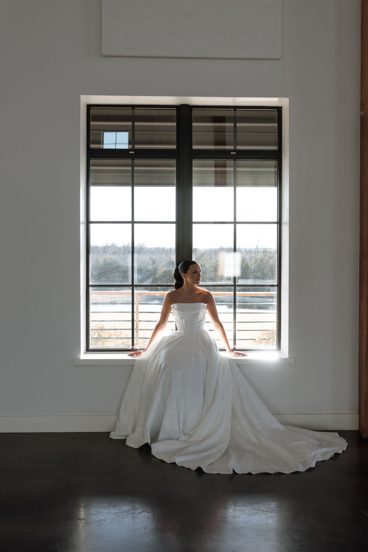 A bride in a flowing white wedding gown sits elegantly by a large window in an empty room.