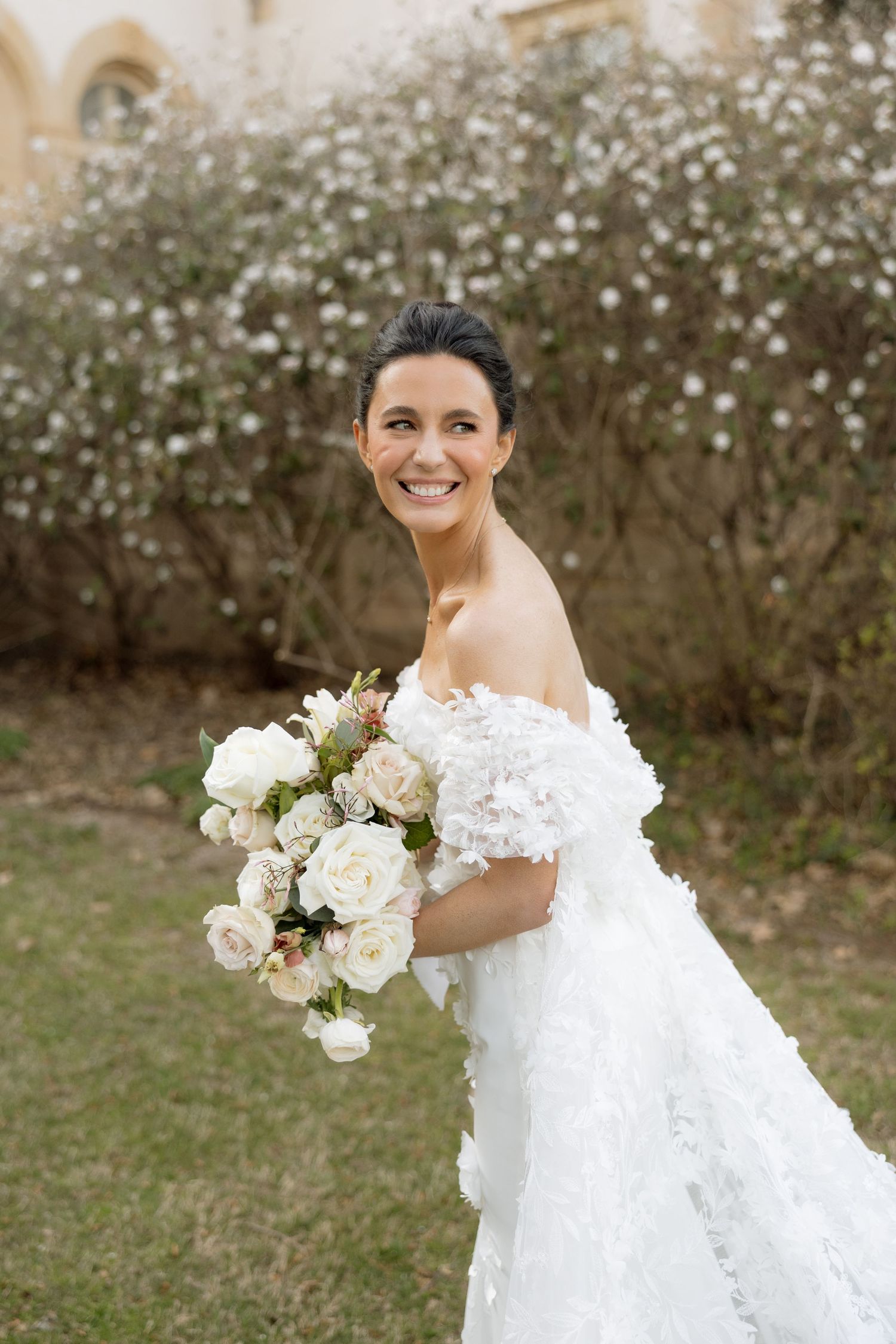 Smiling bride in off-shoulder floral wedding gown holds a lush white rose bouquet in a garden.