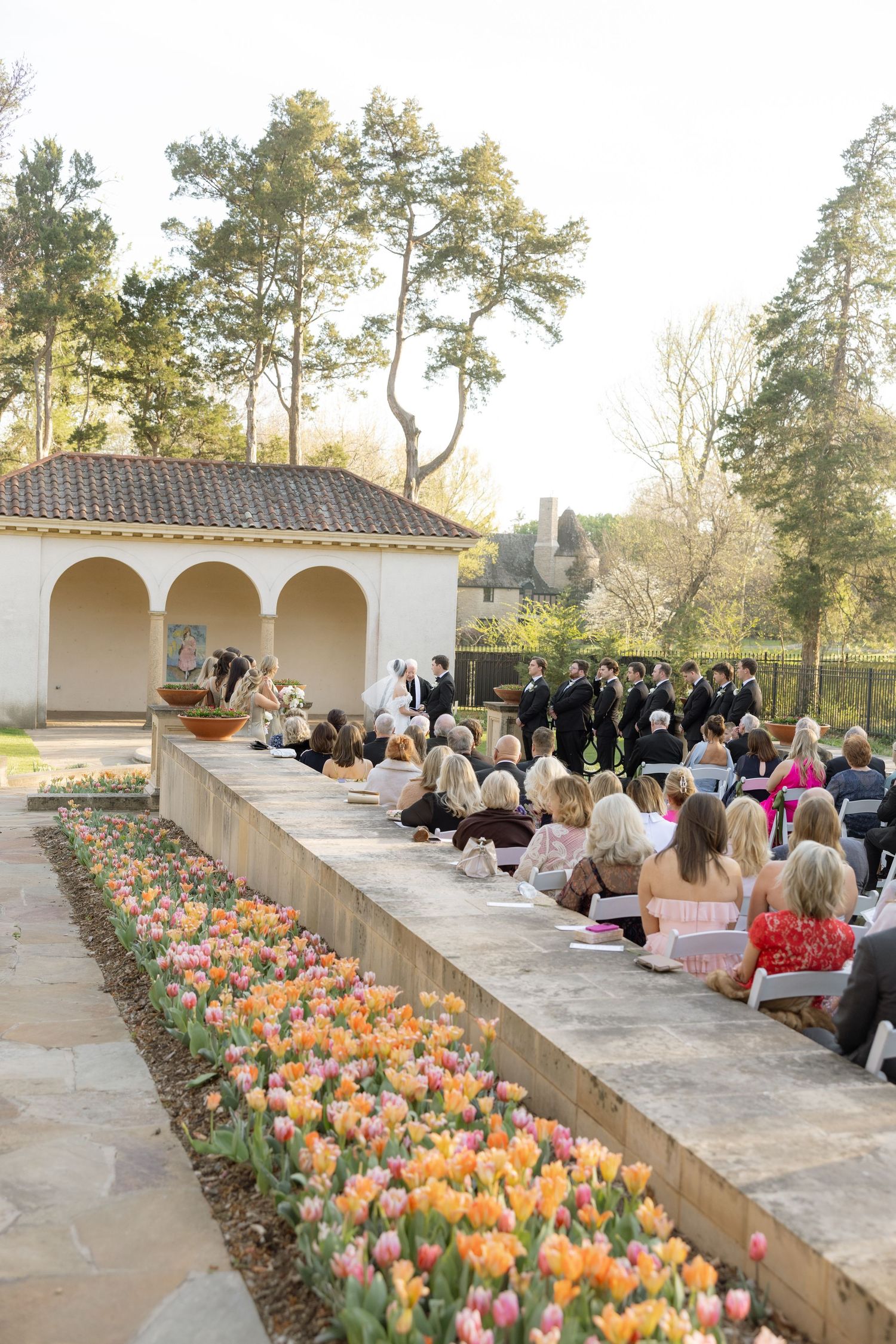 Outdoor wedding ceremony beside colorful tulip garden with guests seated facing altar at elegant estate venue.