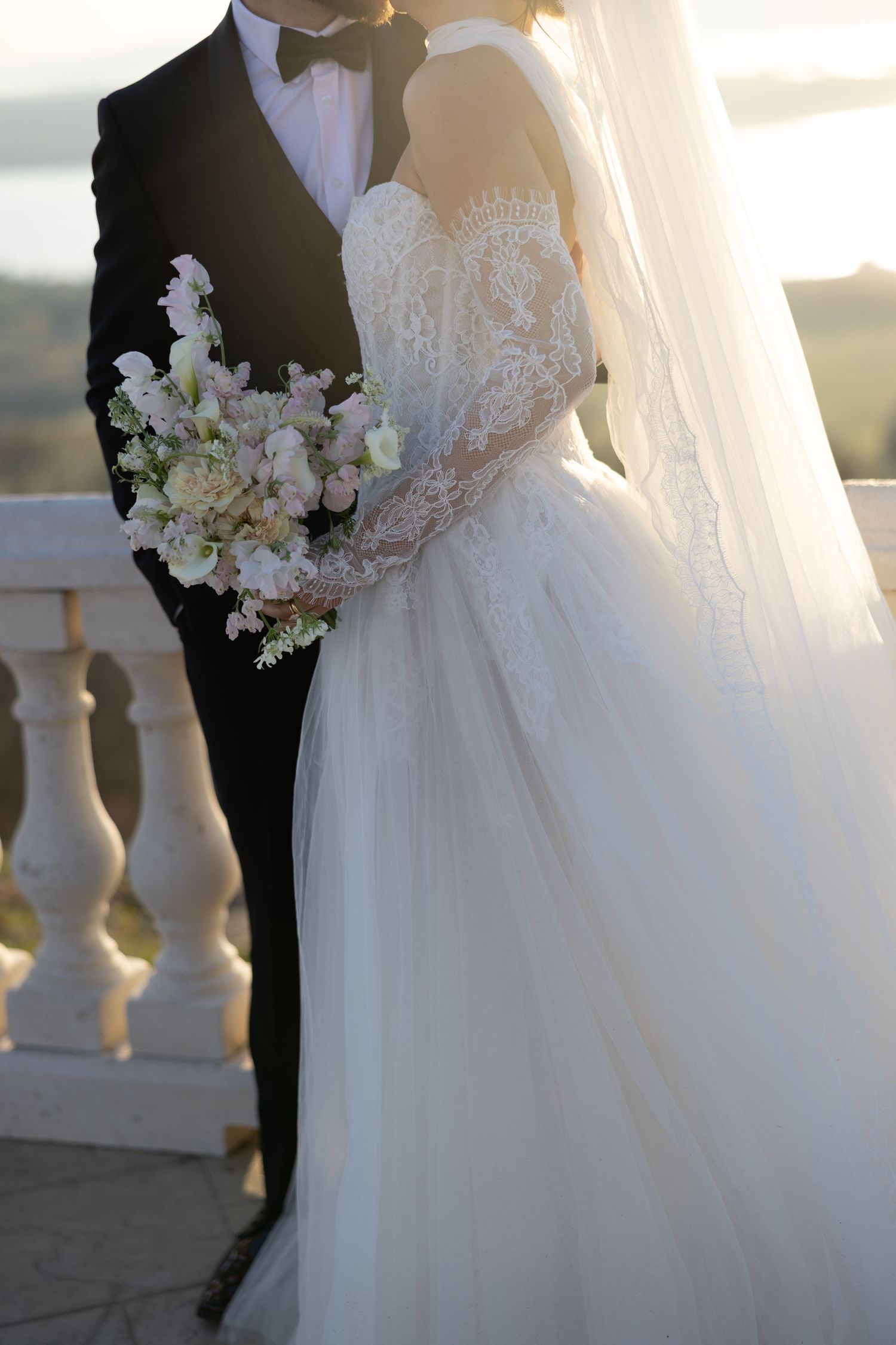 Bride in lace ballgown holding floral bouquet stands with groom in black tuxedo on elegant balcony.