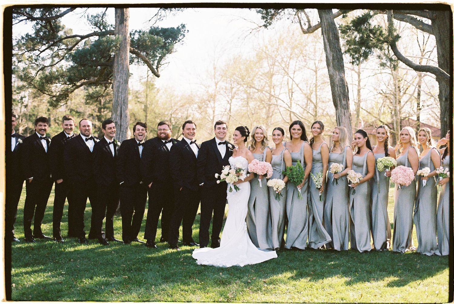 Wedding party poses outdoors with groomsmen in tuxedos and bridesmaids in sage green gowns among trees.