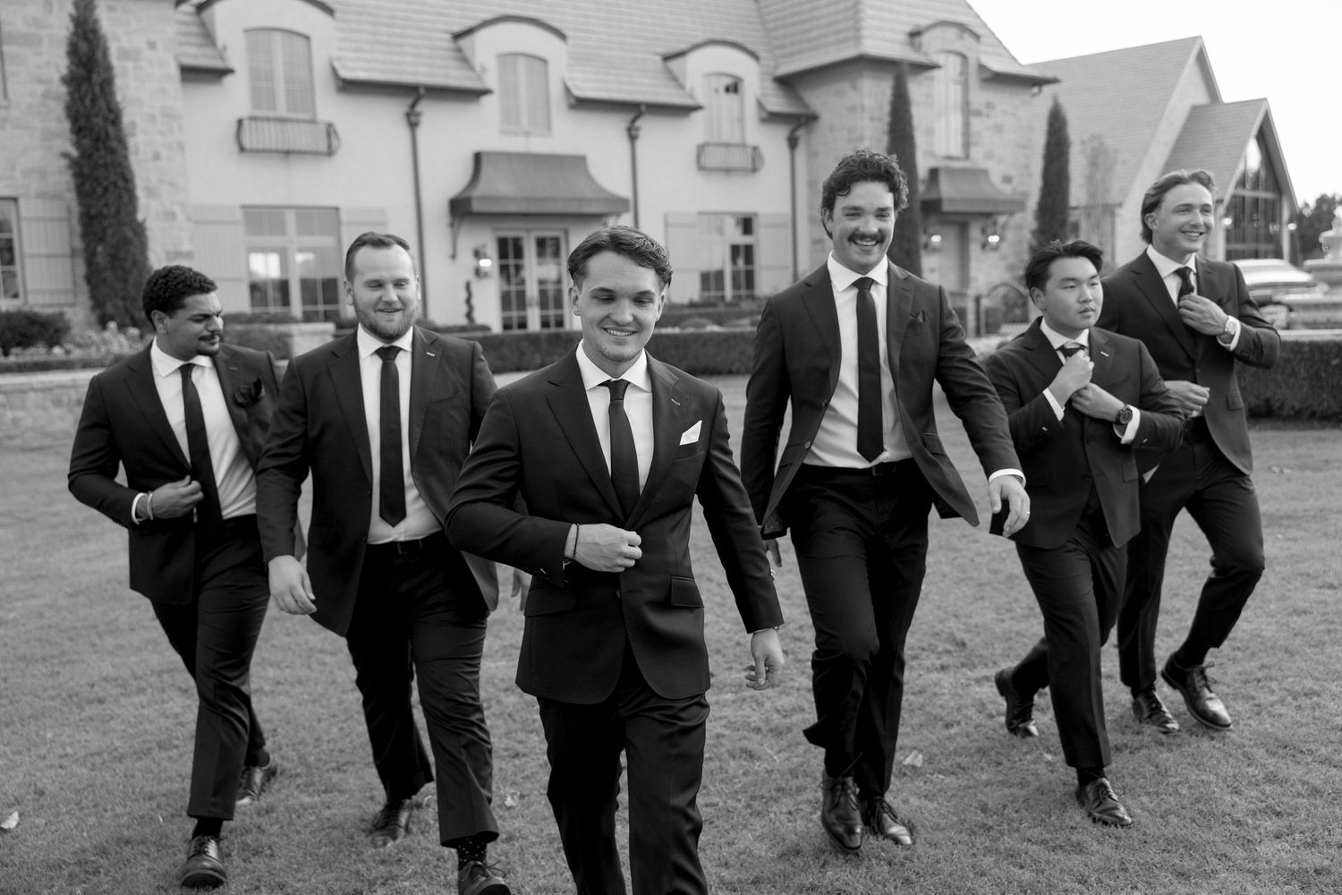 Group of men in dark suits playfully running and jumping outside a stone building.