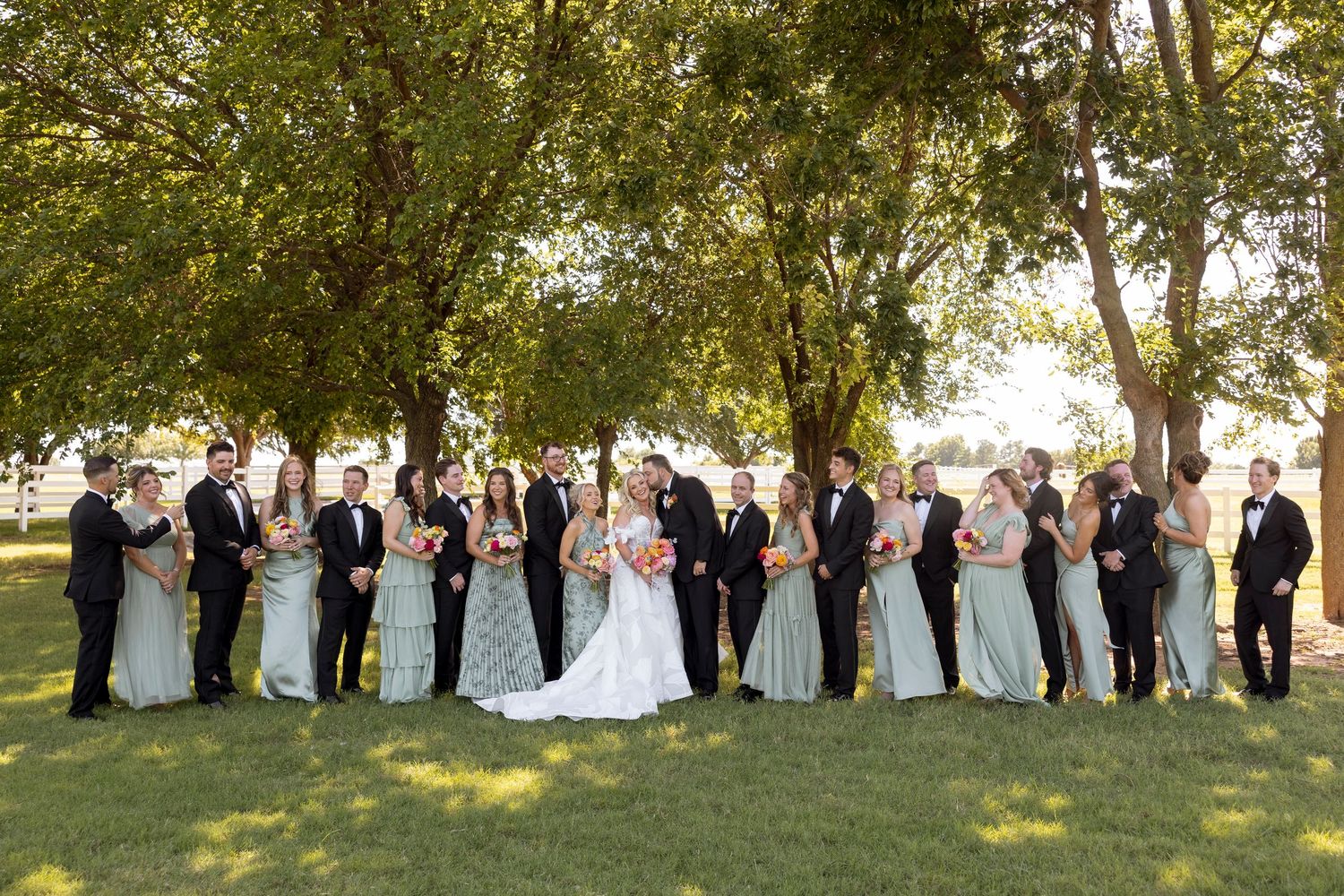 Wedding party lined up outdoors under trees for formal group photos at sunset.