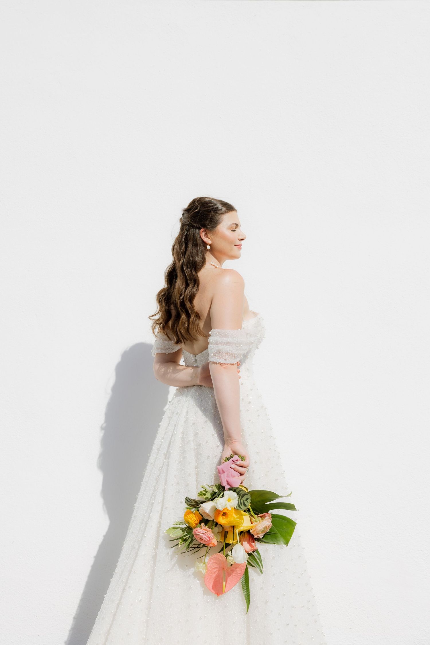 A bride in an off-shoulder lace gown holds a tropical bouquet against a white wall.