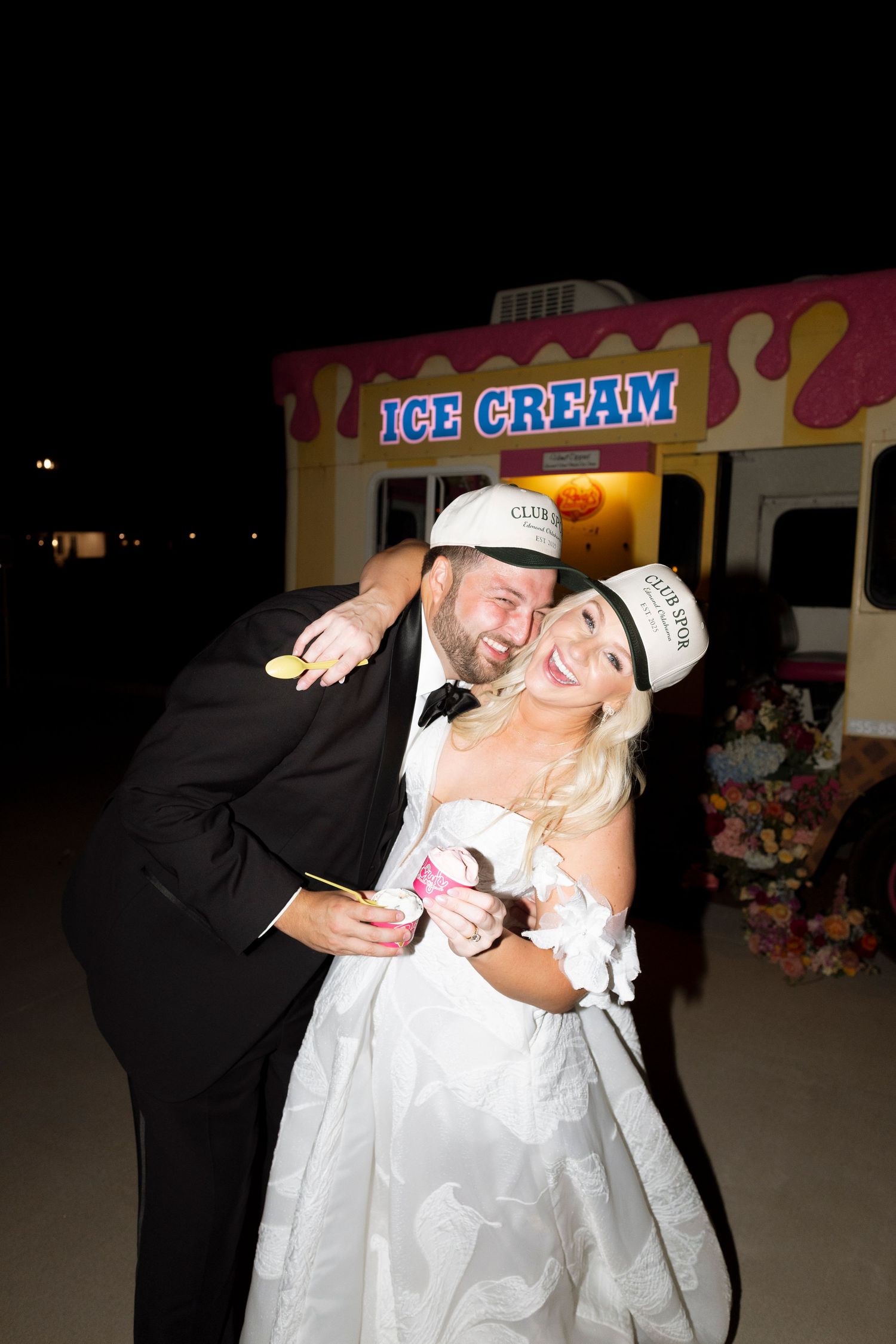 A couple in formal wedding attire share a romantic dance pose at night near an ice cream shop.