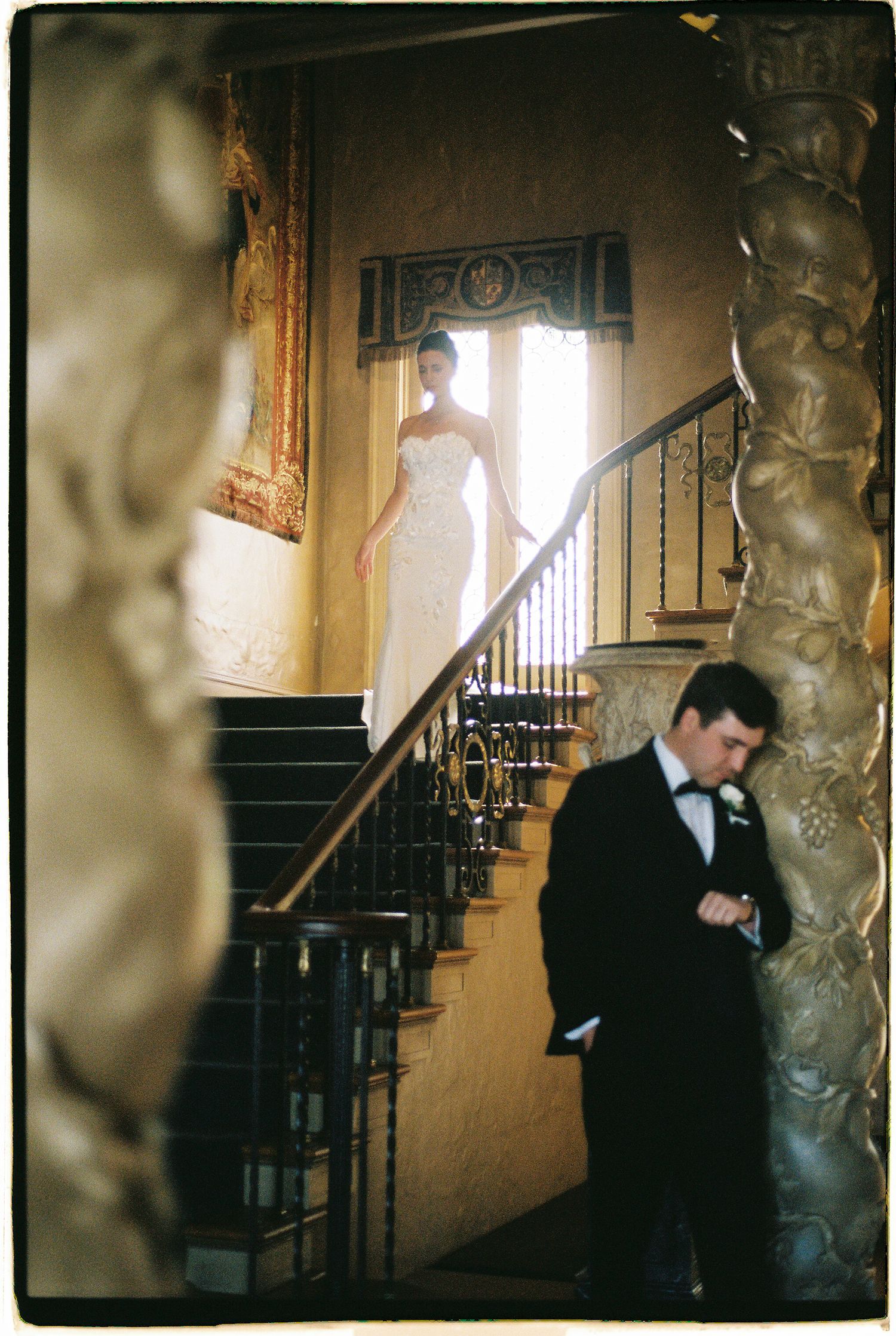 Bride in white gown descends grand staircase while groom in tuxedo waits below at elegant wedding venue.