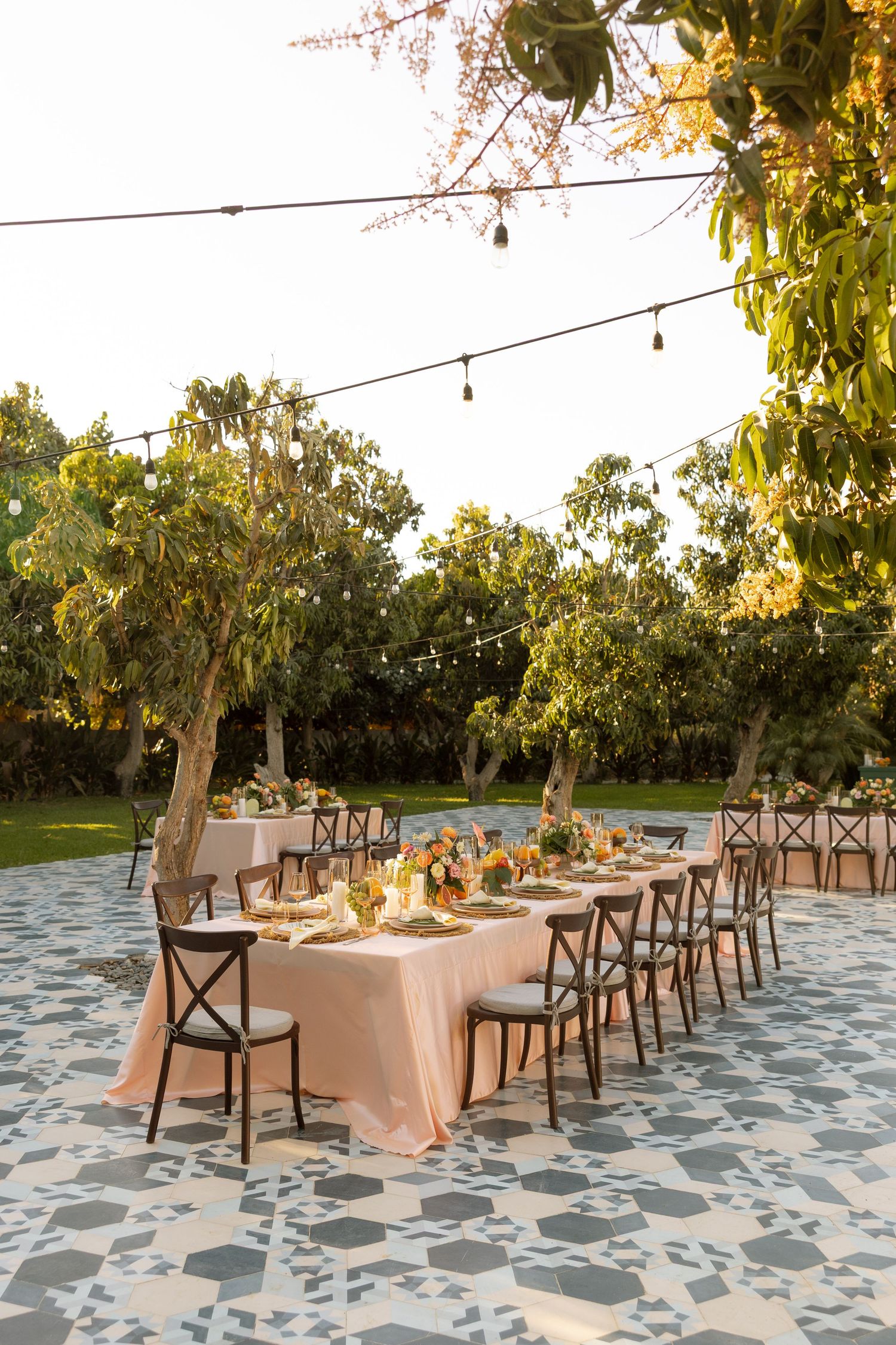 Long outdoor wedding reception table with blush linens under string lights at dusk.