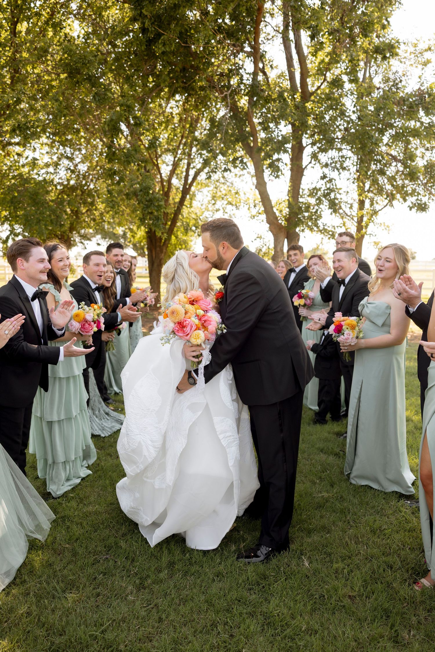 Wedding party in sage green and black attire poses on grassy lawn during golden hour celebrations.