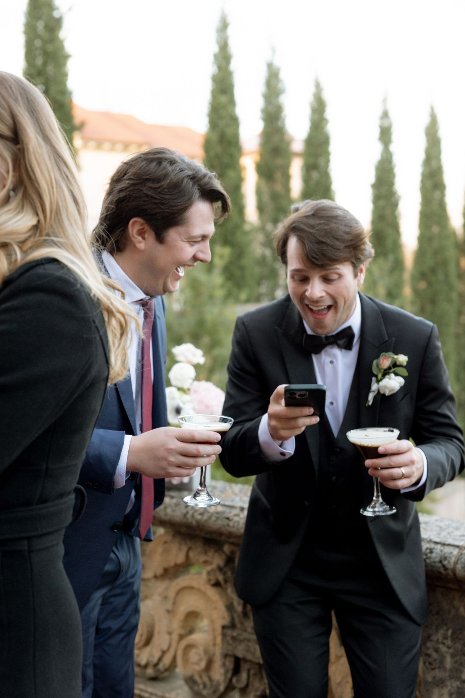 Groom in tuxedo shares phone screen with laughing guest holding cocktails at elegant outdoor wedding reception.